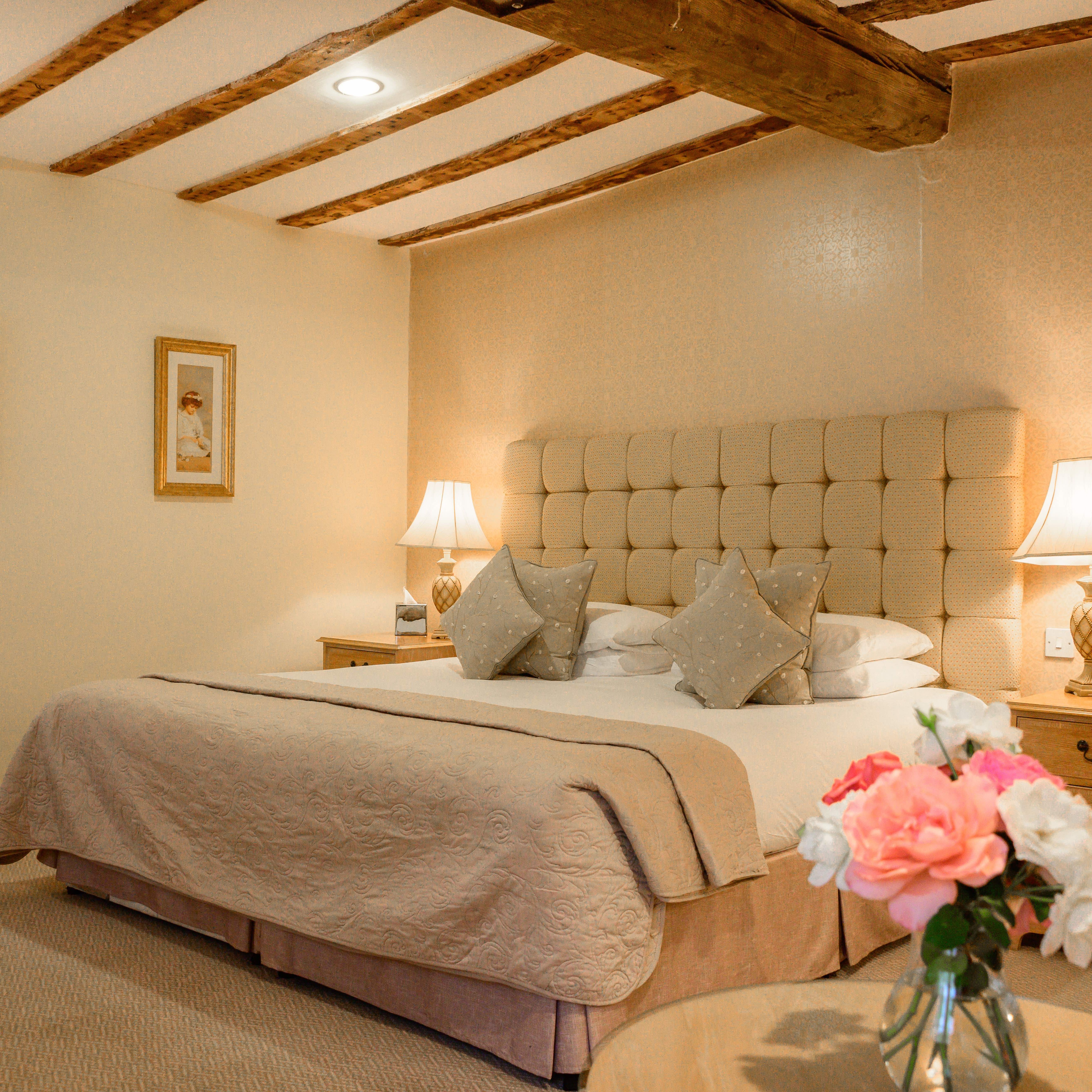 Elegant hotel suite with a large bed, wooden beams on the ceiling, bedside tables with lamps, framed artwork, and a vase of flowers on the table.