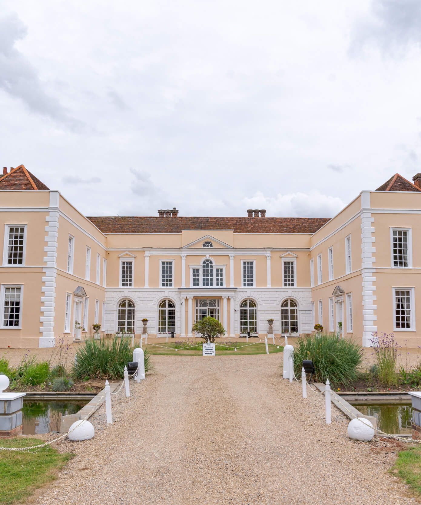 Large historic manor house with symmetrical architecture, cream-colored walls, and a central gravel pathway leading to the entrance.