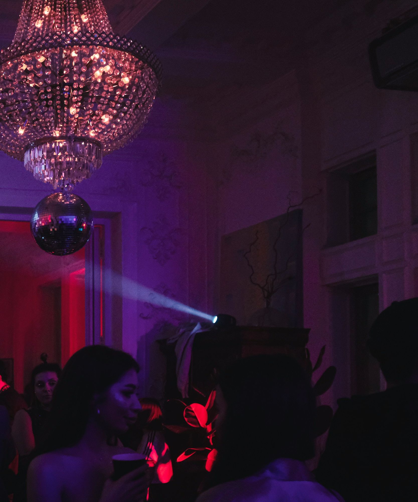 A dimly lit party scene with people socializing under a sparkling chandelier and a disco ball, illuminated by colorful lights.