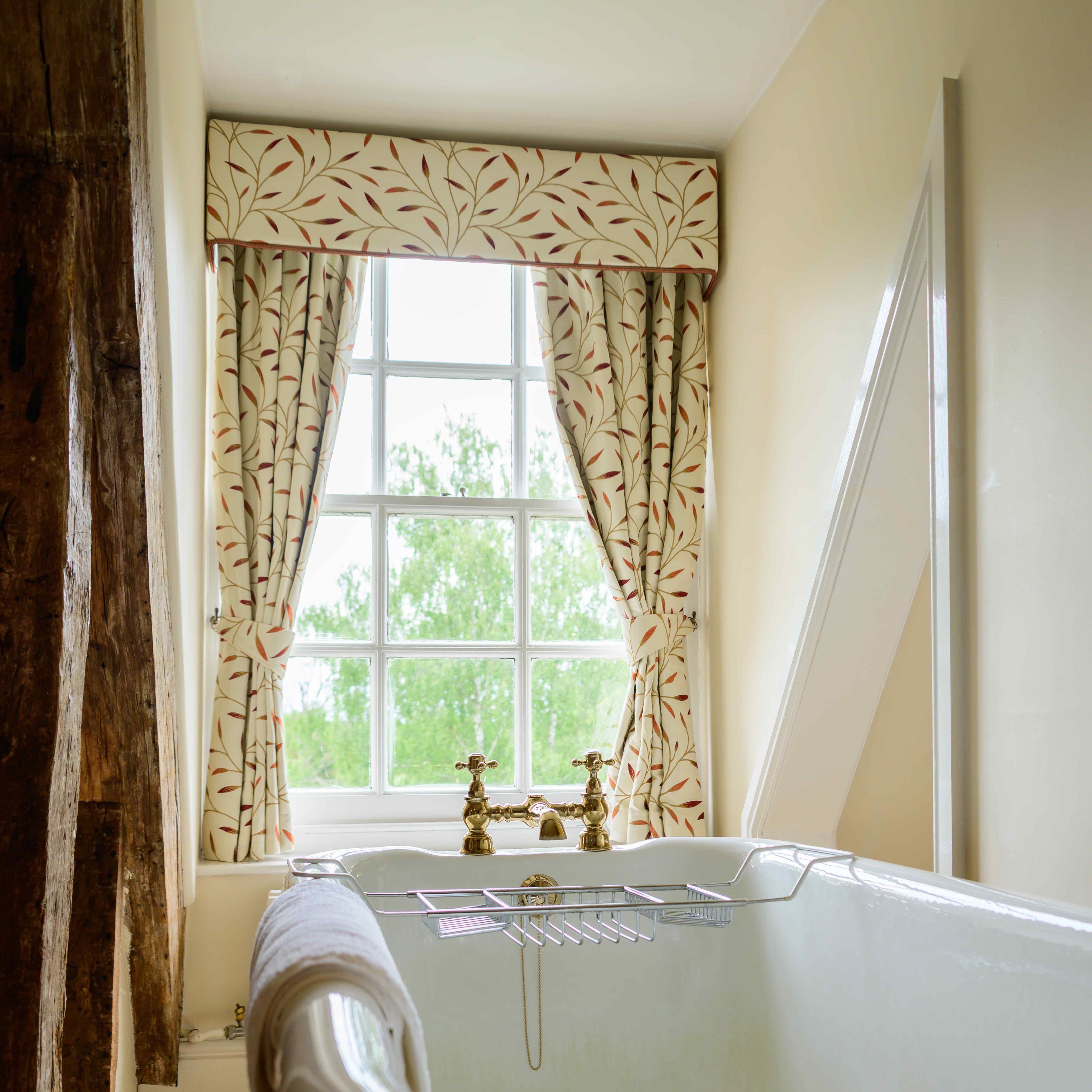 Freestanding bathtub in a bathroom with a window, patterned curtains, and vintage brass taps.