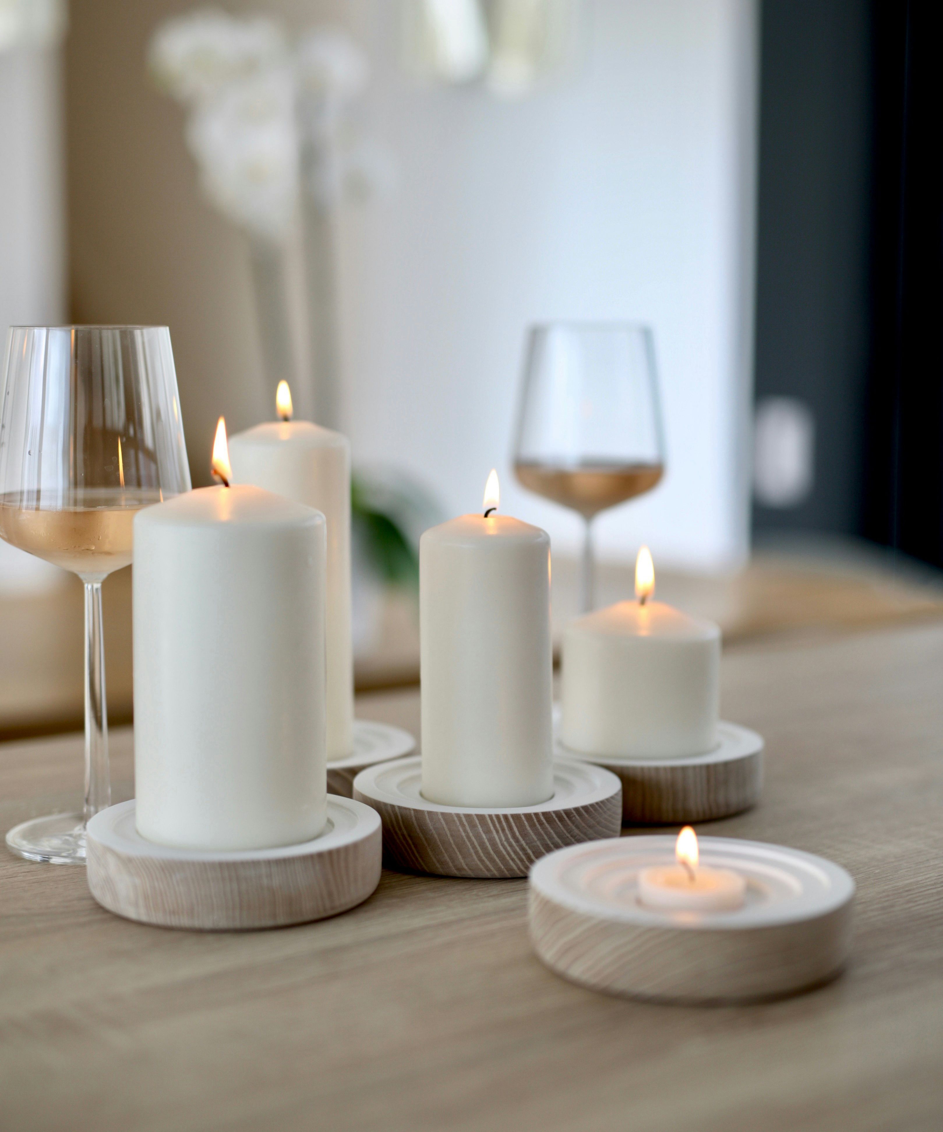 Lit white candles of varying heights on wooden holders next to wine glasses on a wooden table.