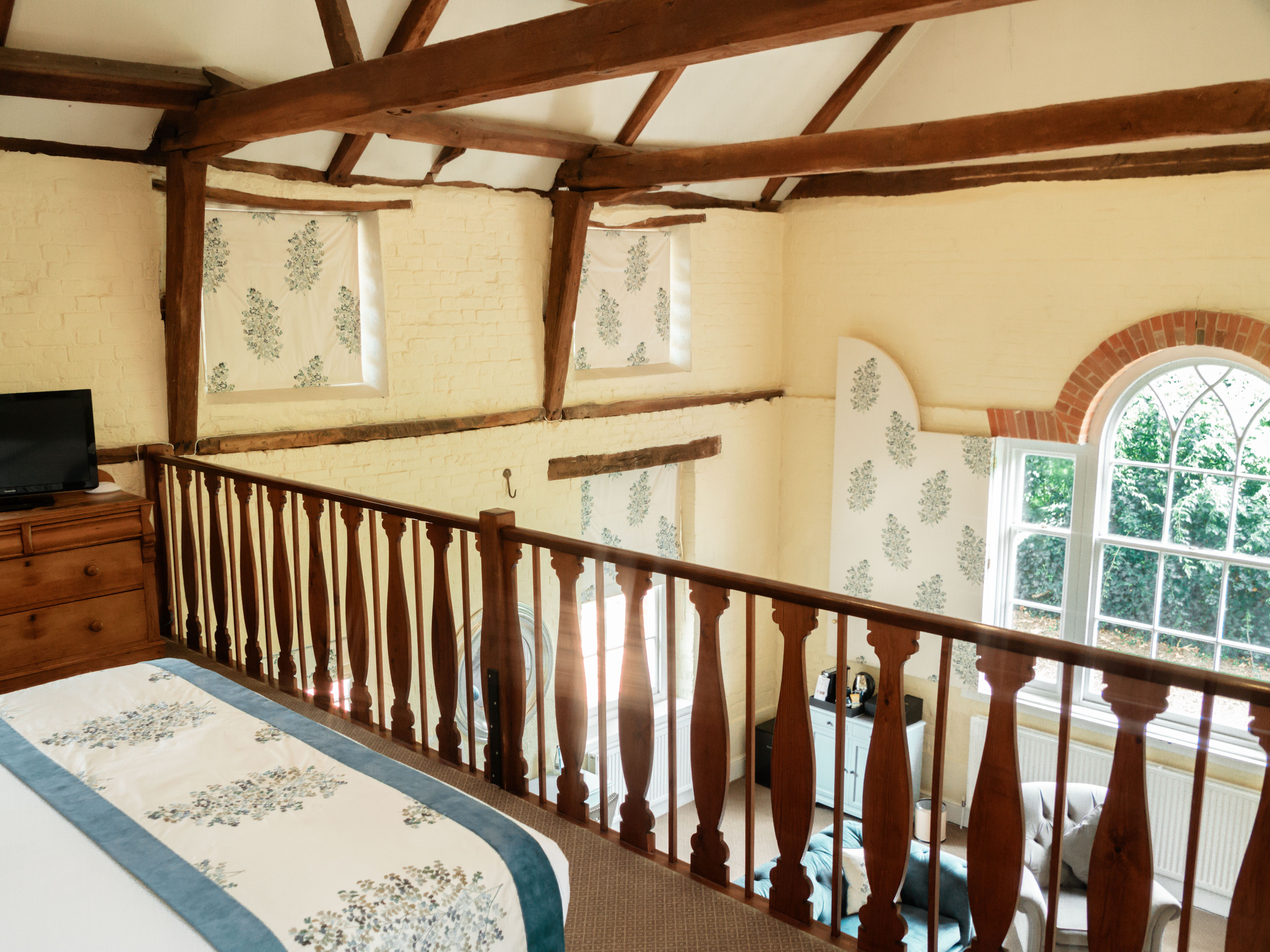 Loft bedroom with wooden beams, floral decor, and a view of a cozy sitting area through a large arched window.