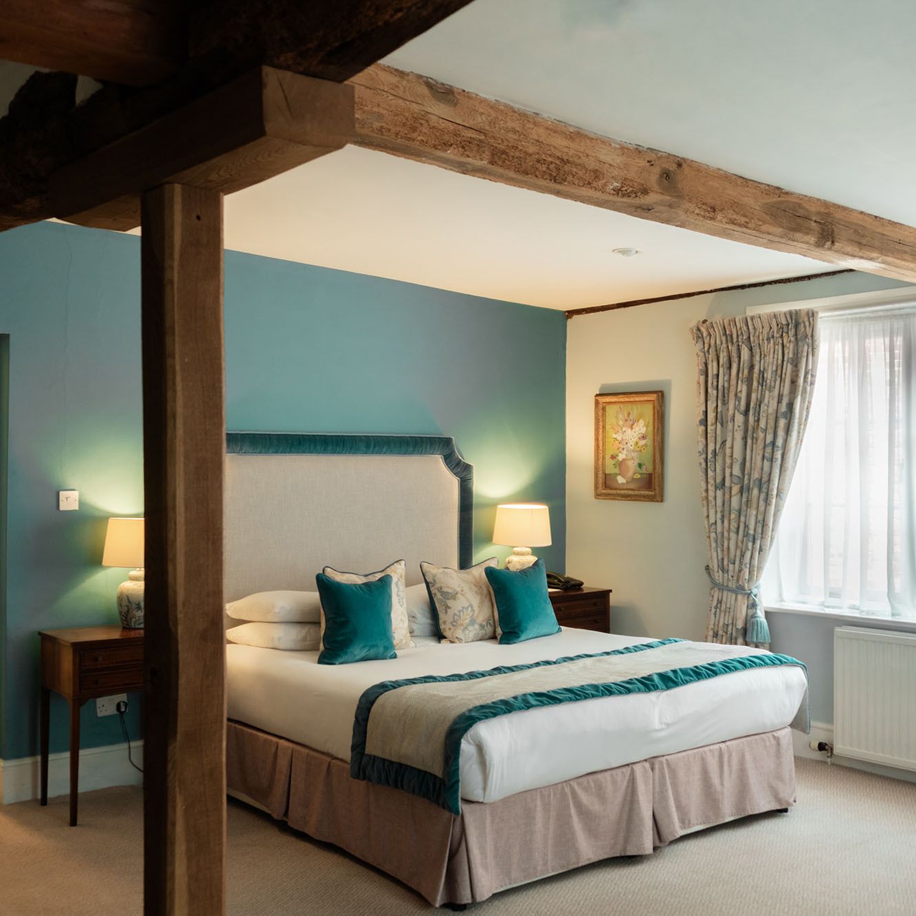 Cozy hotel room with exposed wooden beams, a large bed, teal pillows, and an ensuite bathroom.
