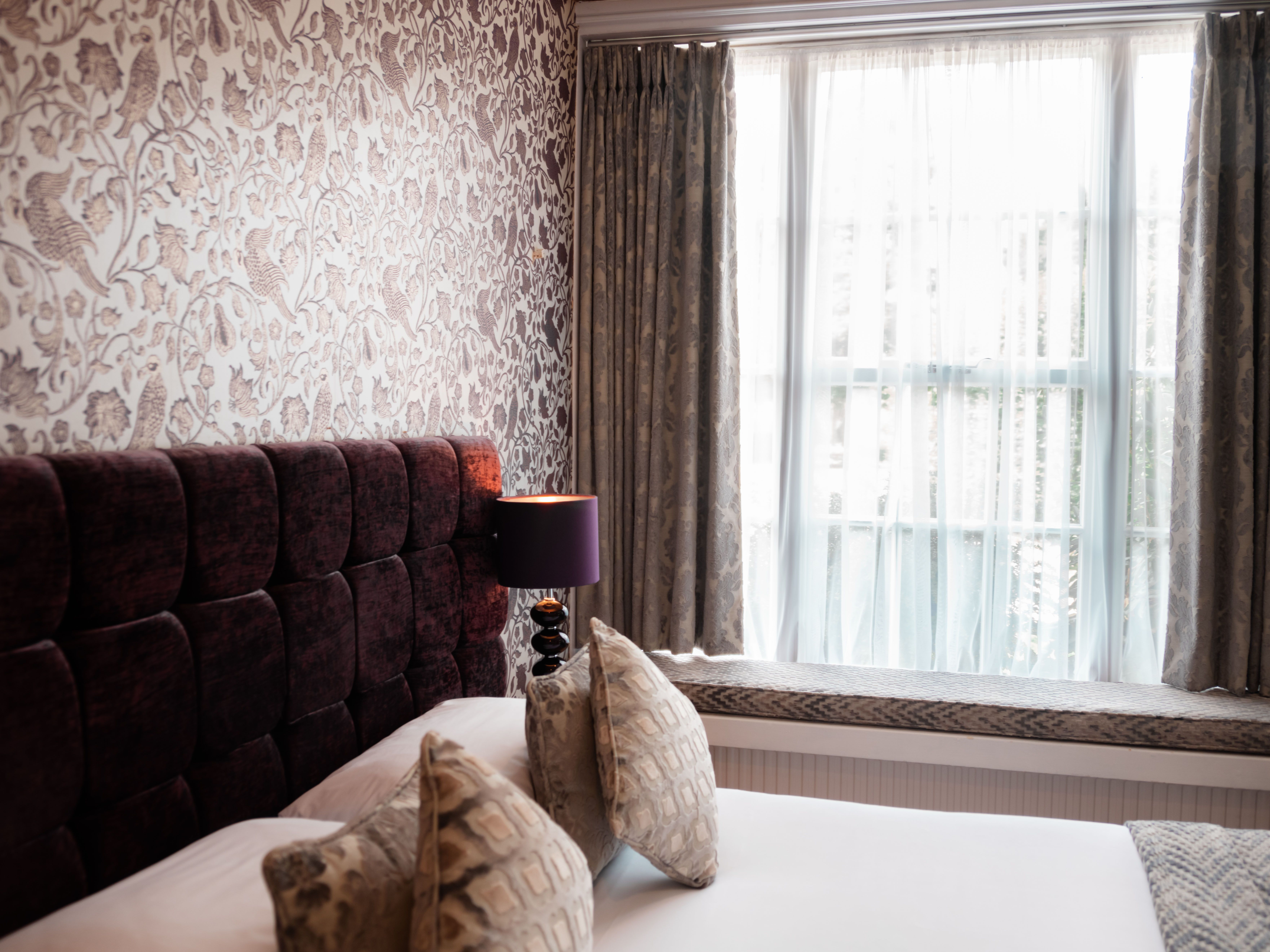 Cozy bedroom with patterned wallpaper, a padded headboard, and large window with curtains