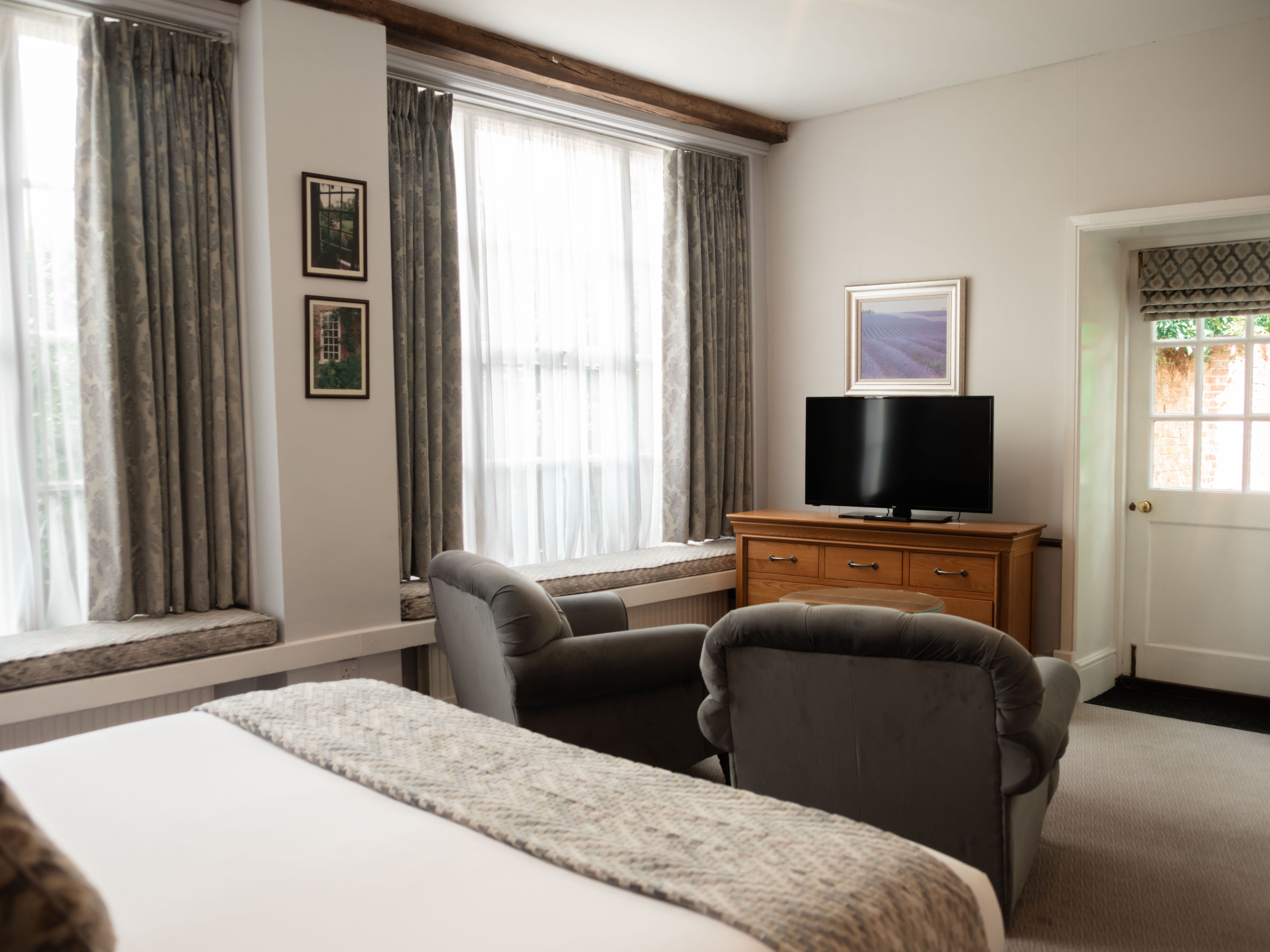 Cozy hotel room with double bed, two armchairs, dresser with TV, and large windows with curtains.