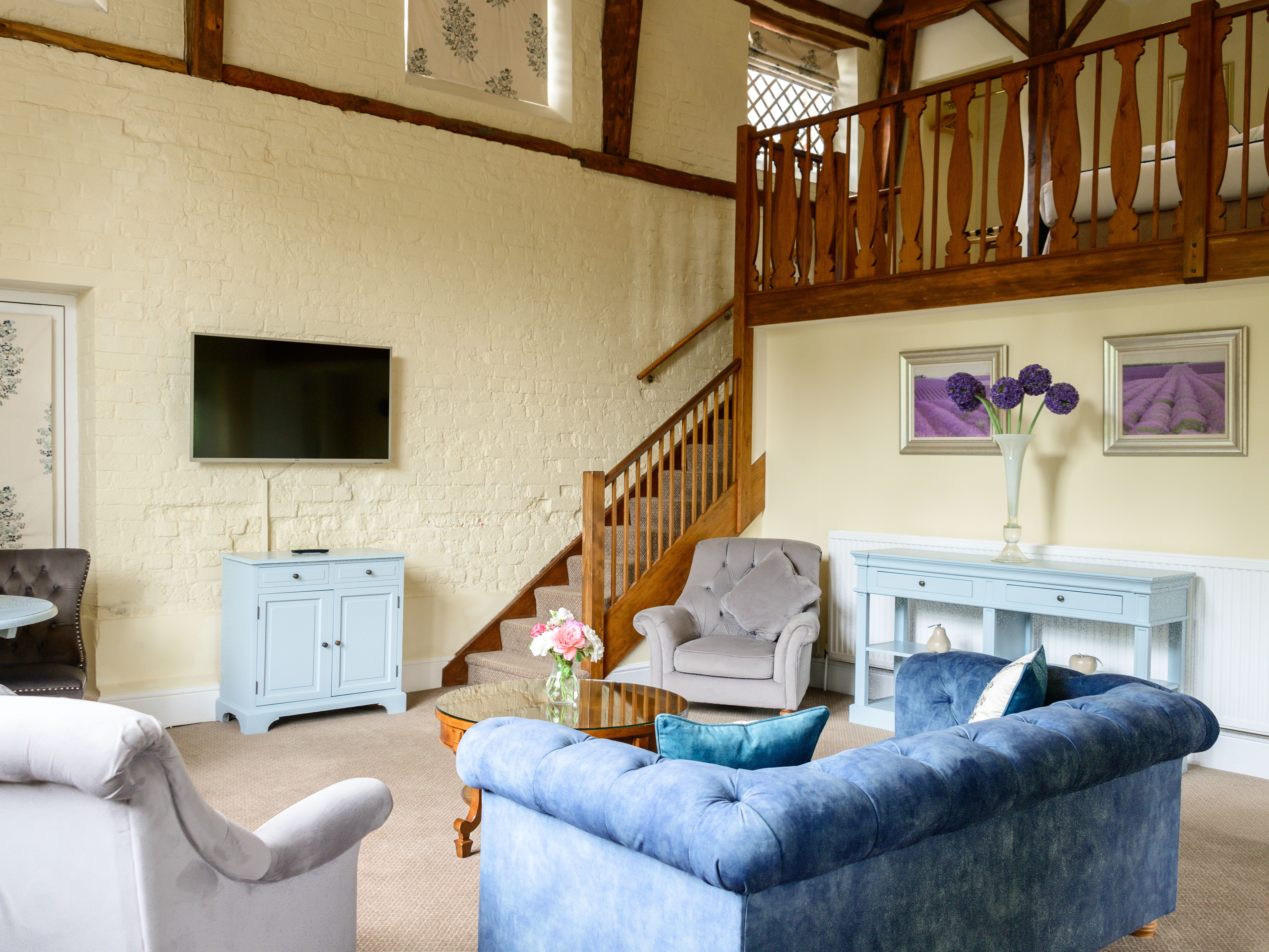 Cozy living room with blue and grey furniture, light yellow walls, a TV mounted on the wall, and a wooden staircase leading to a loft.