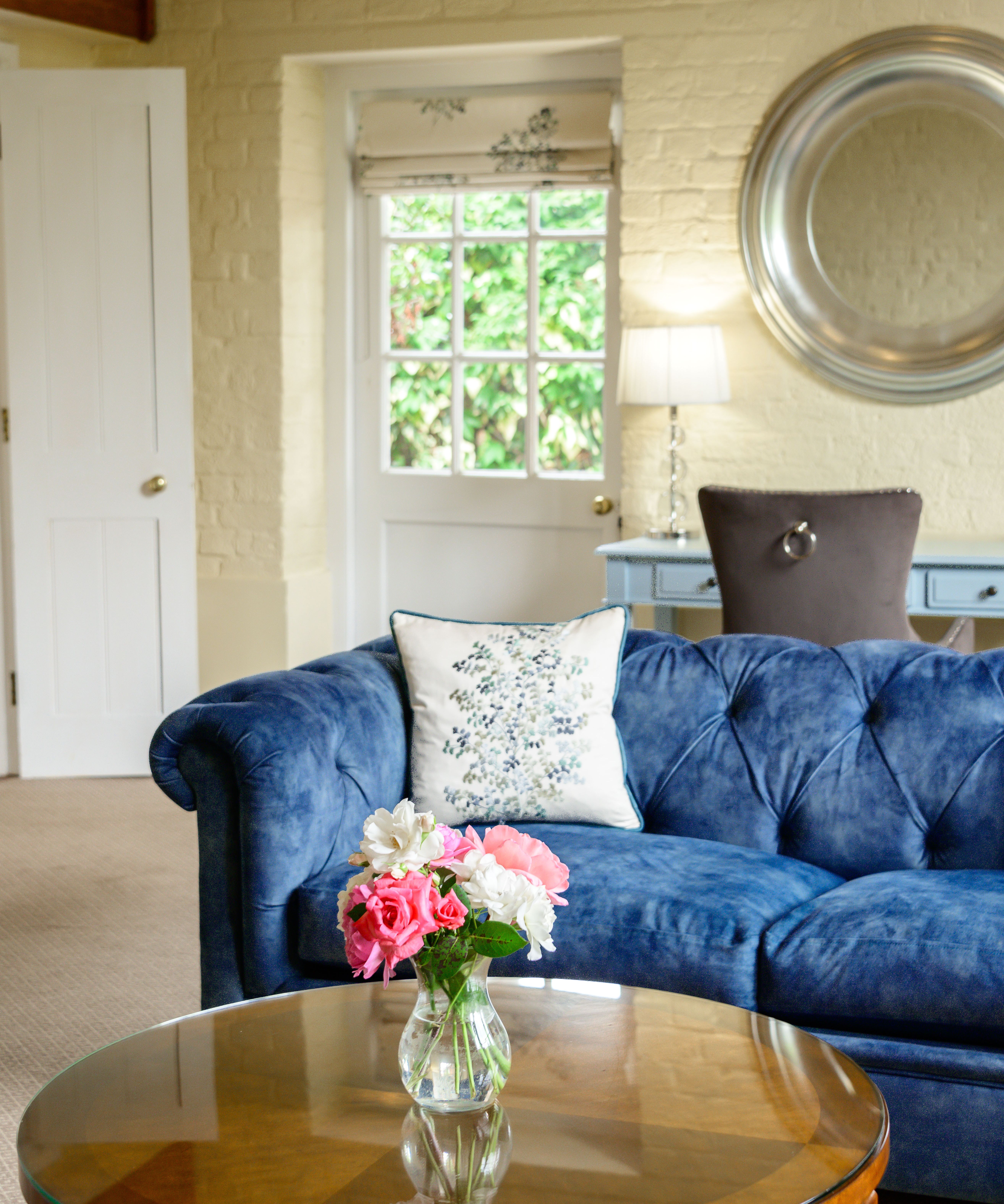 Living room with a blue velvet sofa, floral pillow, round wooden coffee table with a vase of flowers, and a window in the background.