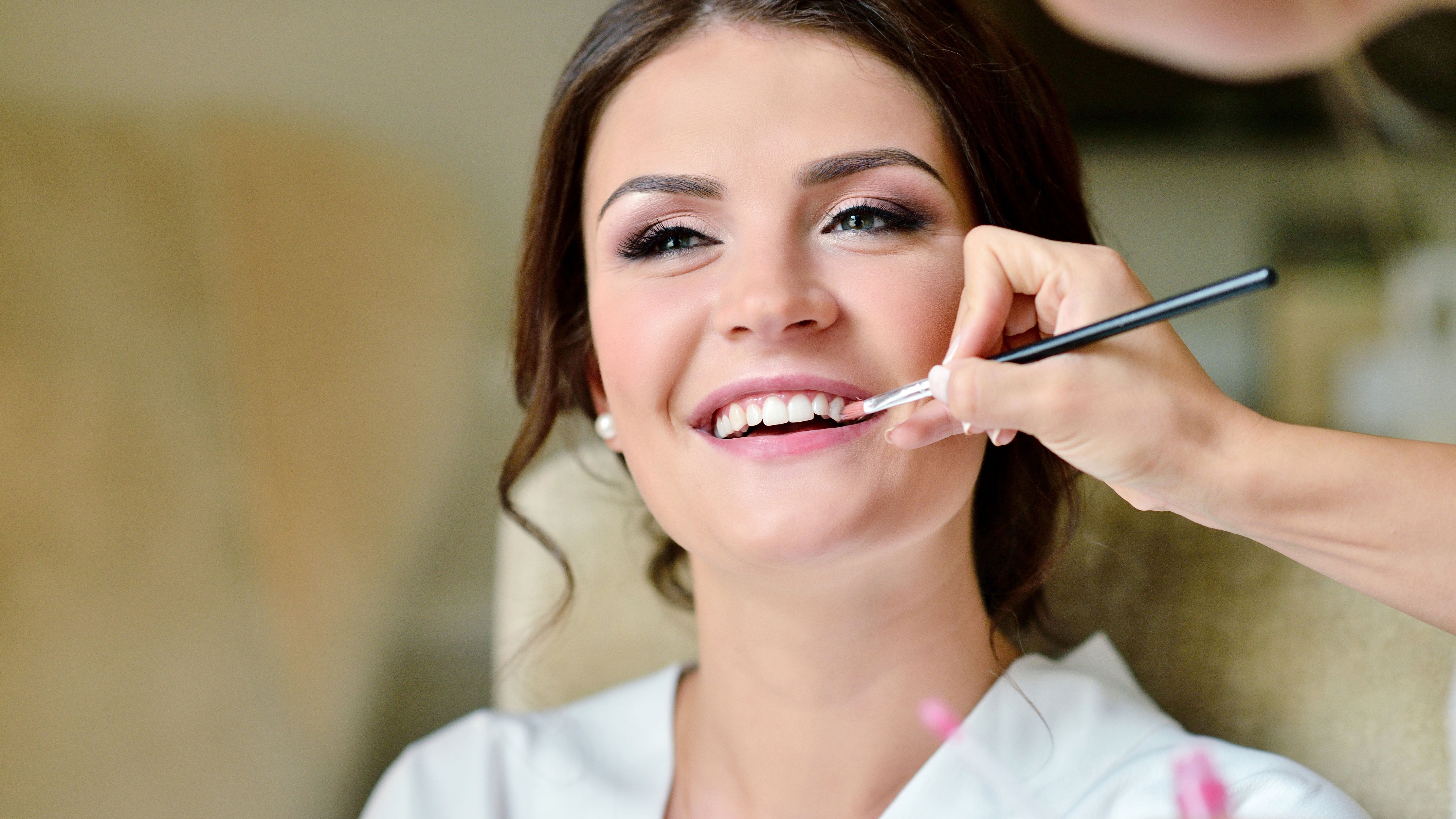 Woman smiling while getting her makeup done, with a makeup artist applying lipstick using a brush.
