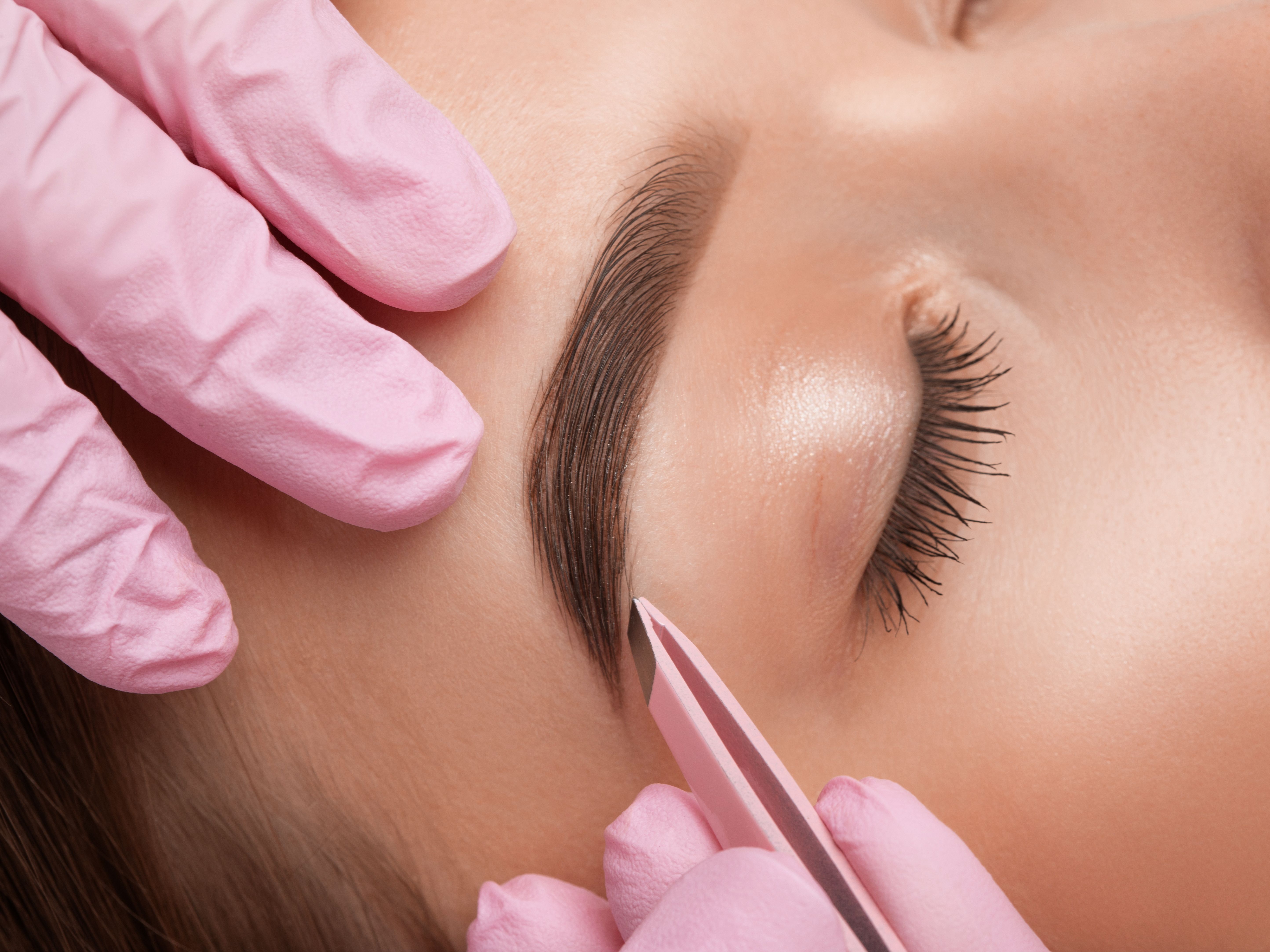 Close-up of a person having their eyebrows shaped with tweezers by a professional wearing pink gloves.