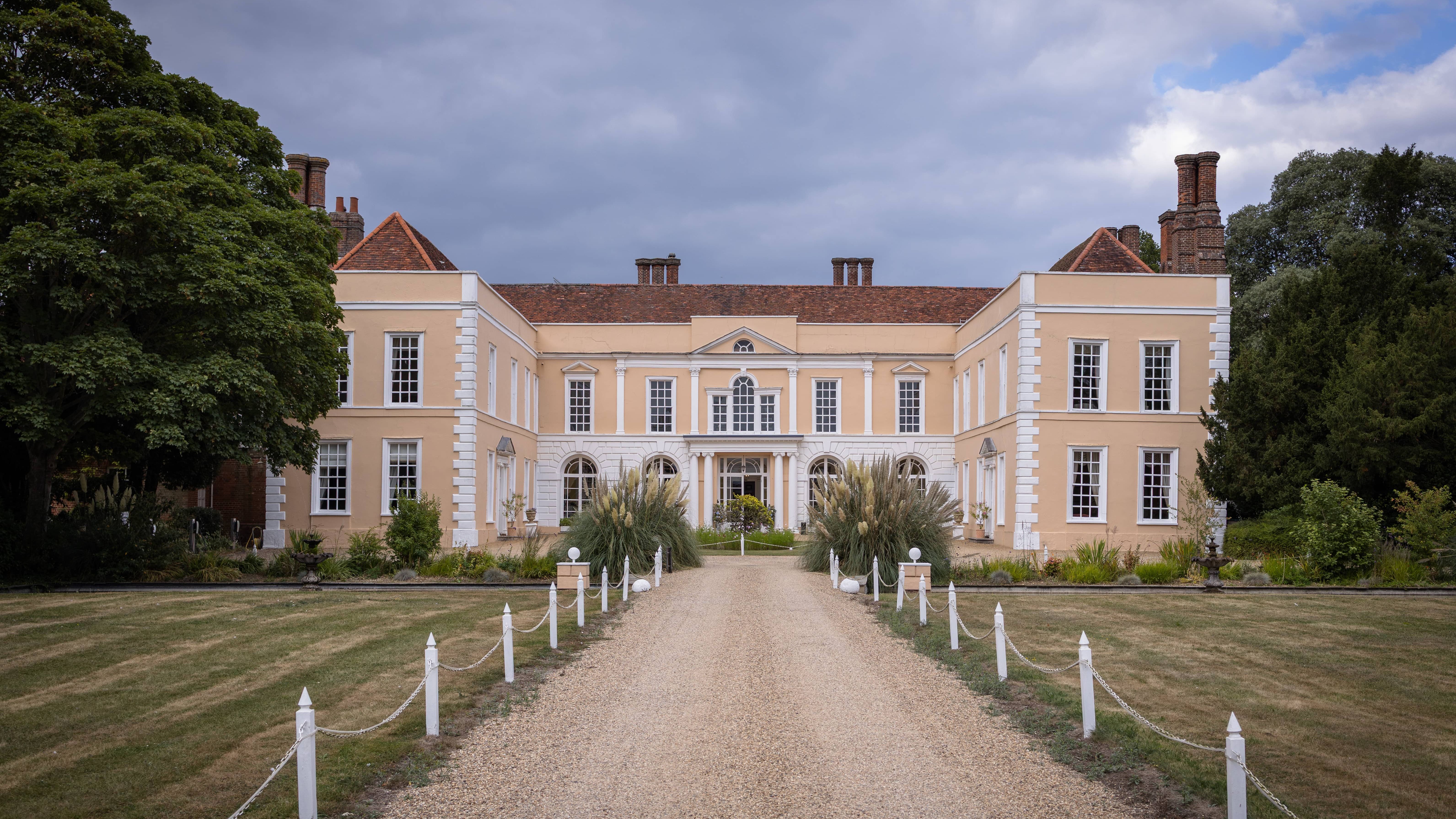 A large, classical mansion with beige walls and red-tiled roof, surrounded by greenery and a gravel driveway leading to the entrance.