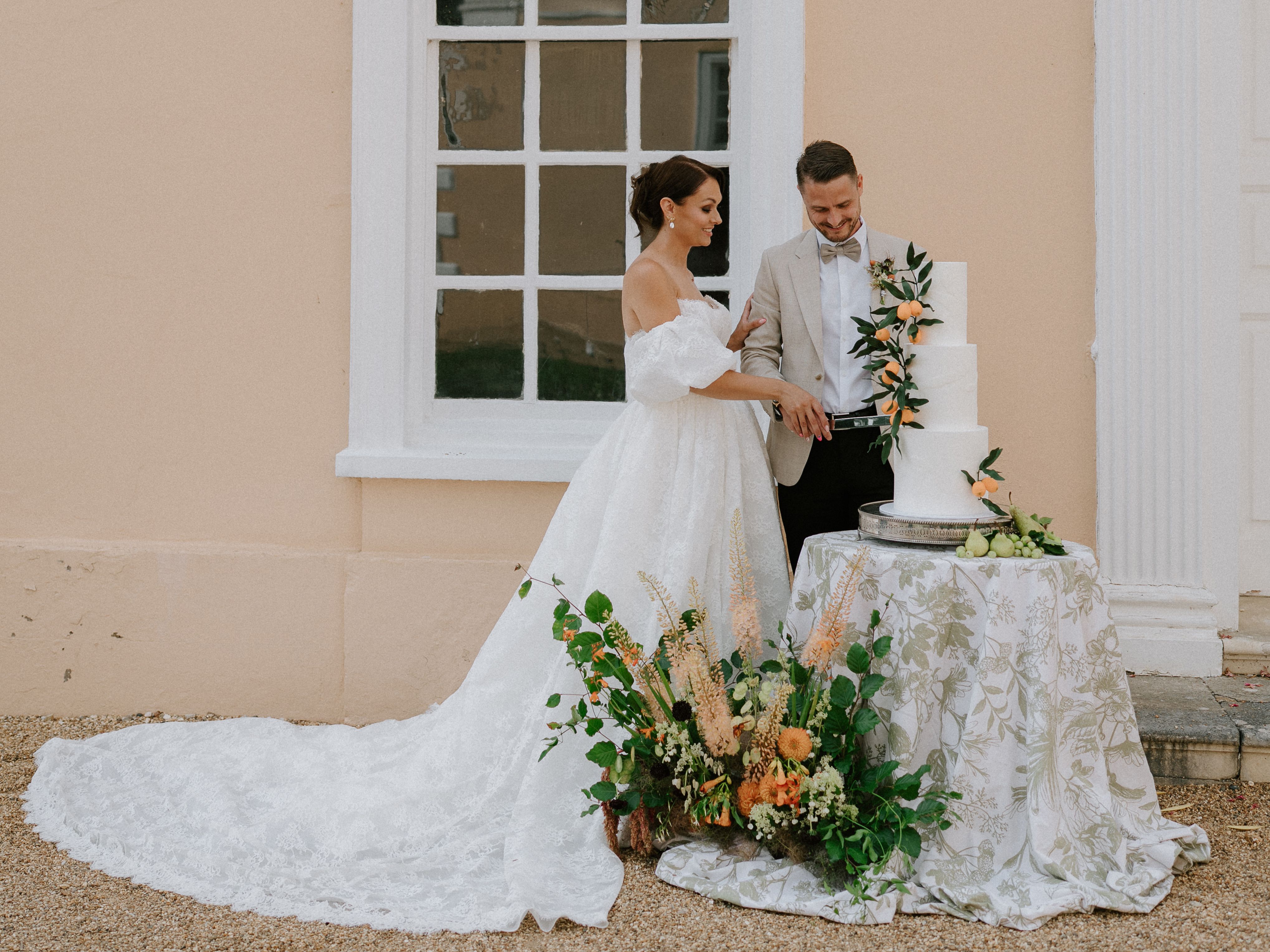 Bride and groom cutting a wedding cake outdoors beside a pastel-colored building.