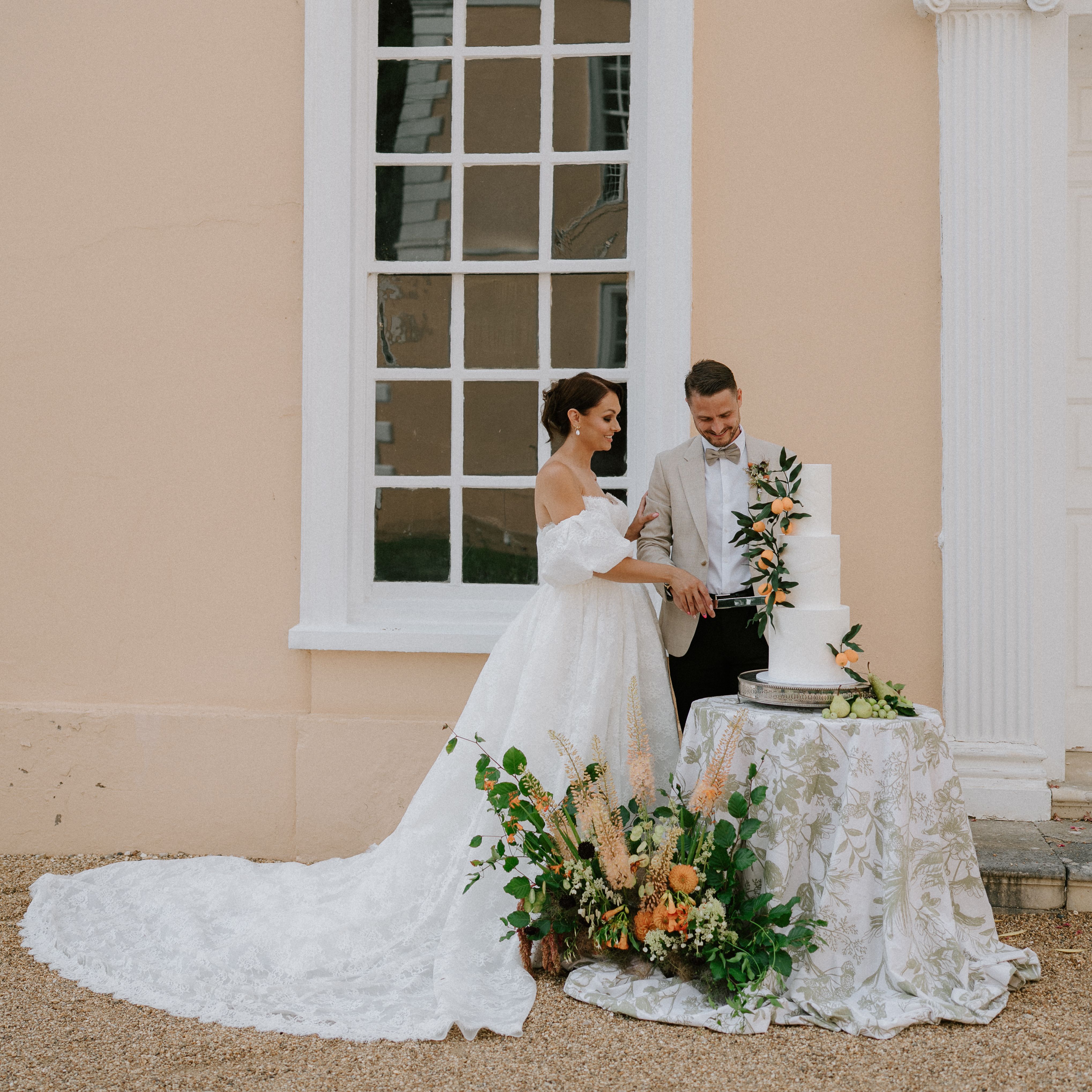 Bride and groom cutting a wedding cake outdoors beside a pastel-colored building.