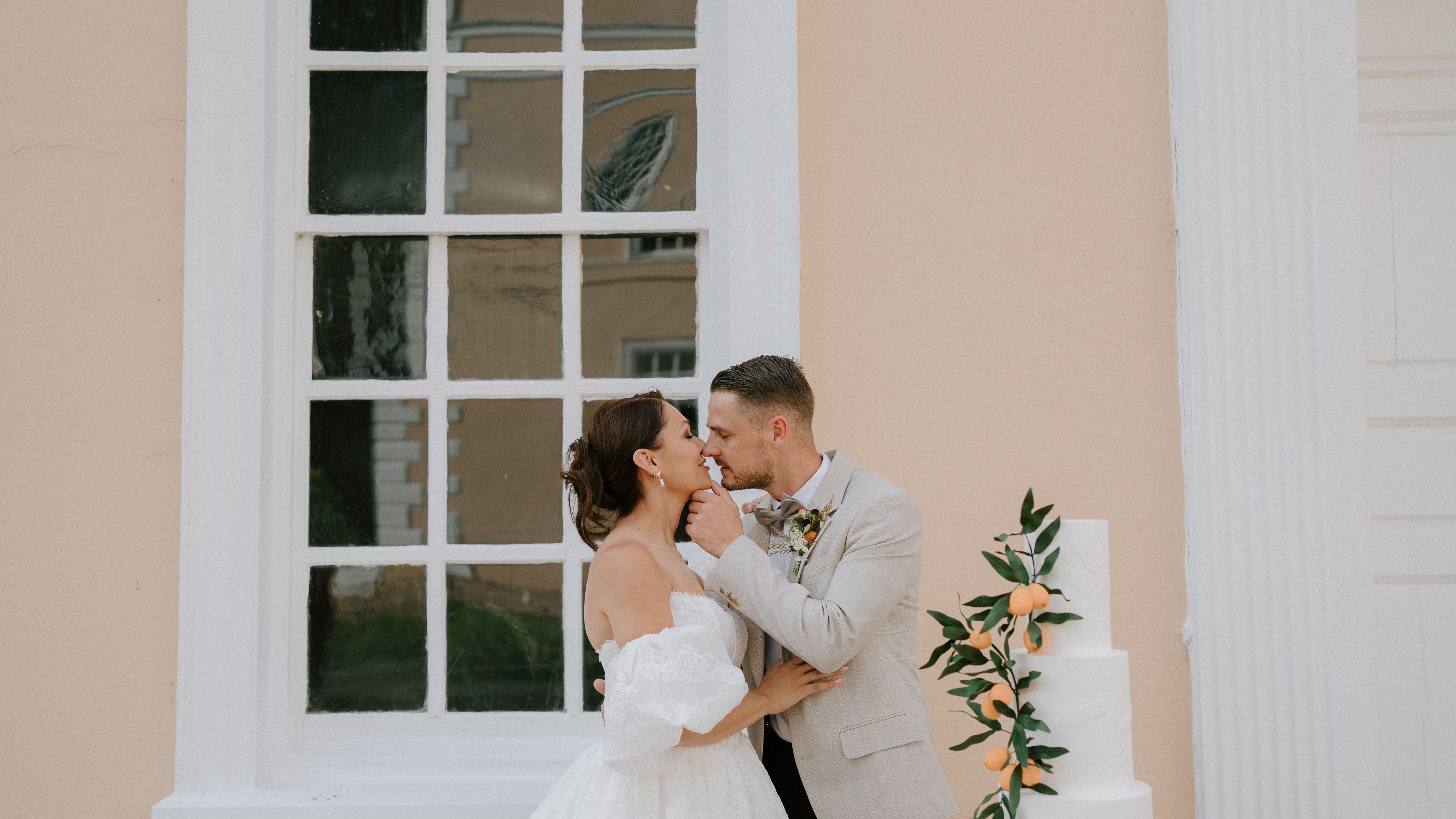 Bride and groom embracing next to a white wedding cake decorated with oranges and greenery in front of a large window.