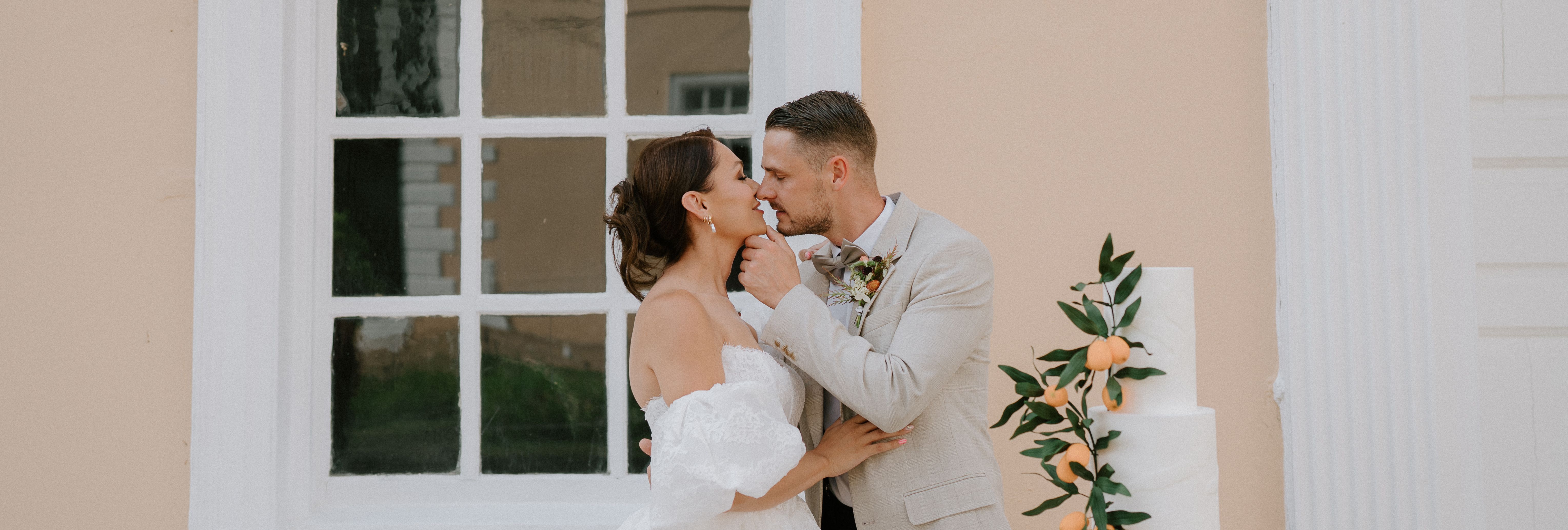 Bride and groom embracing next to a white wedding cake decorated with oranges and greenery in front of a large window.