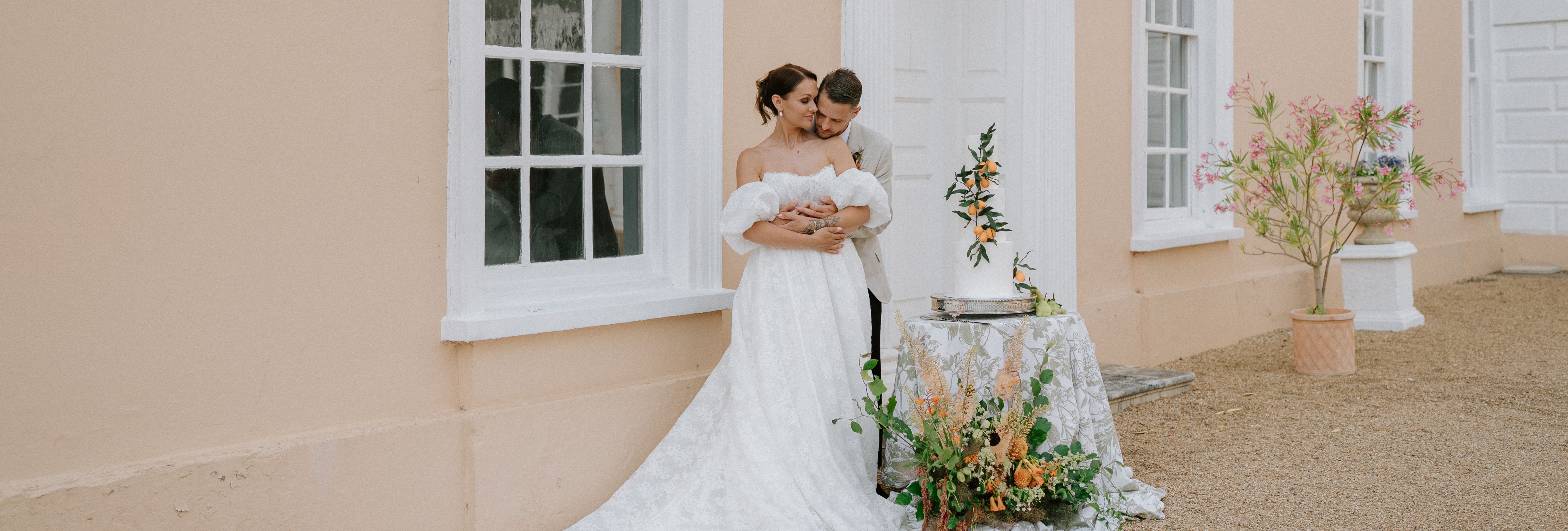 Bride and groom embracing outside a peach-colored building with wedding cake and floral arrangements.
