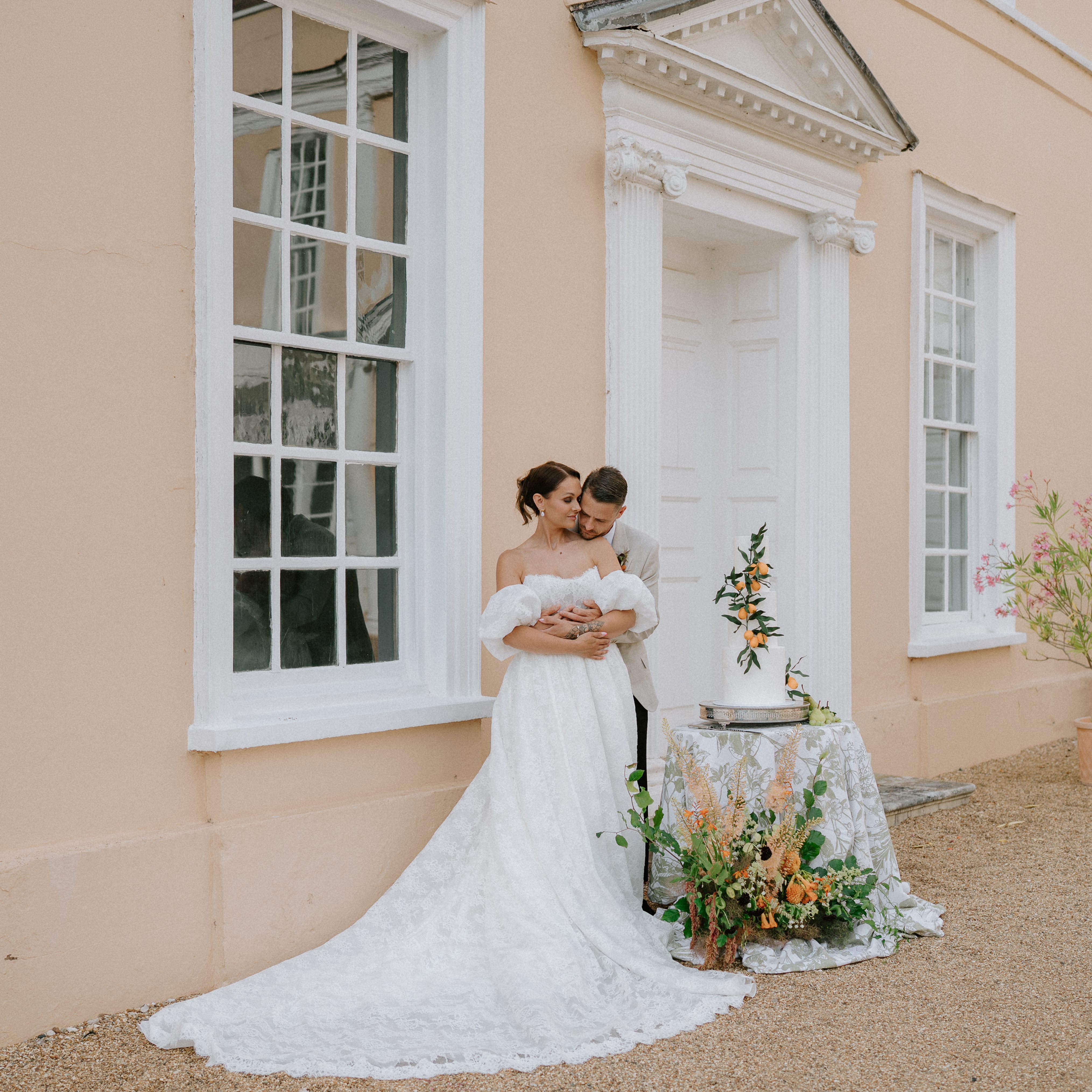 Bride and groom embracing outside a peach-colored building with wedding cake and floral arrangements.