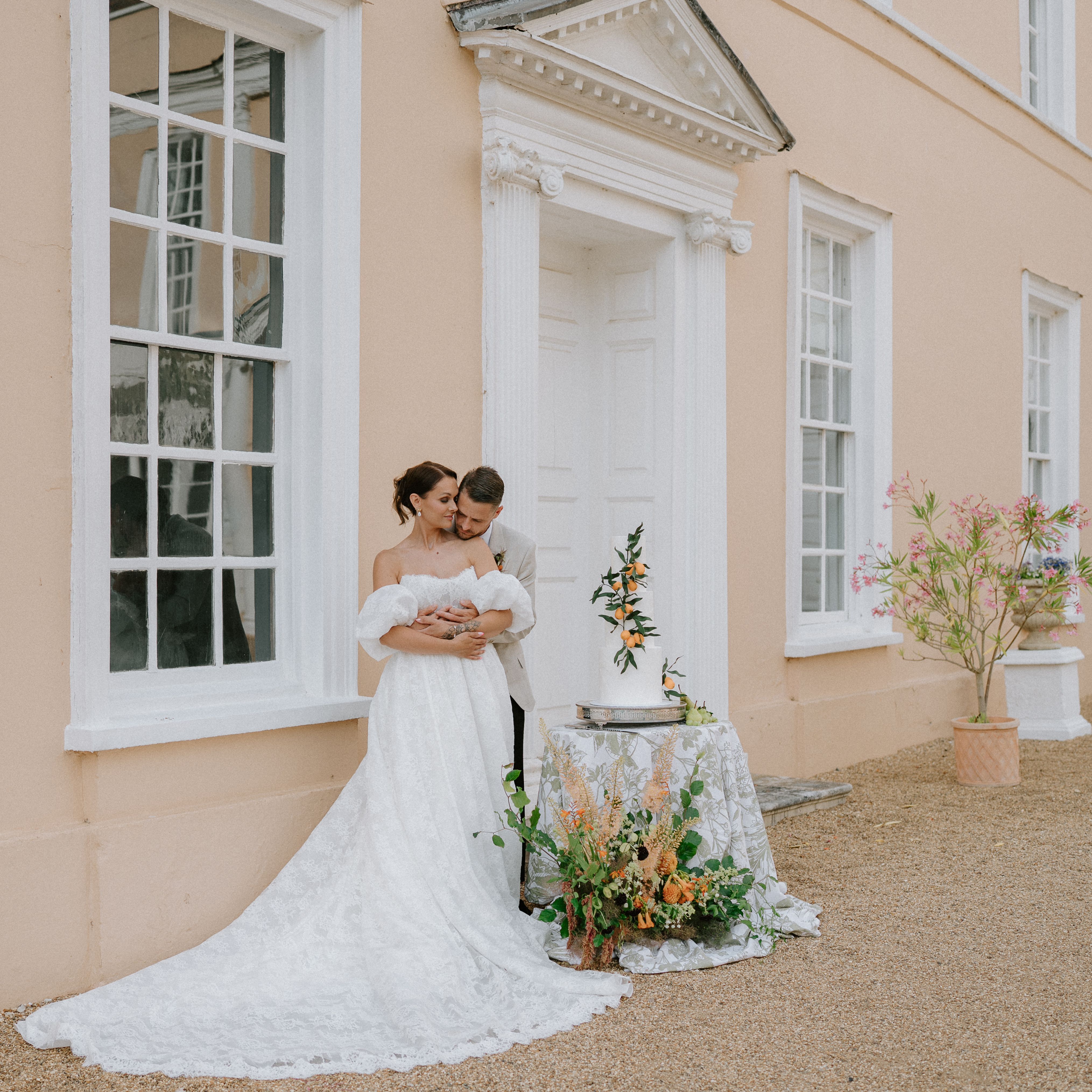 Bride and groom embracing outside a peach-colored building with wedding cake and floral arrangements.