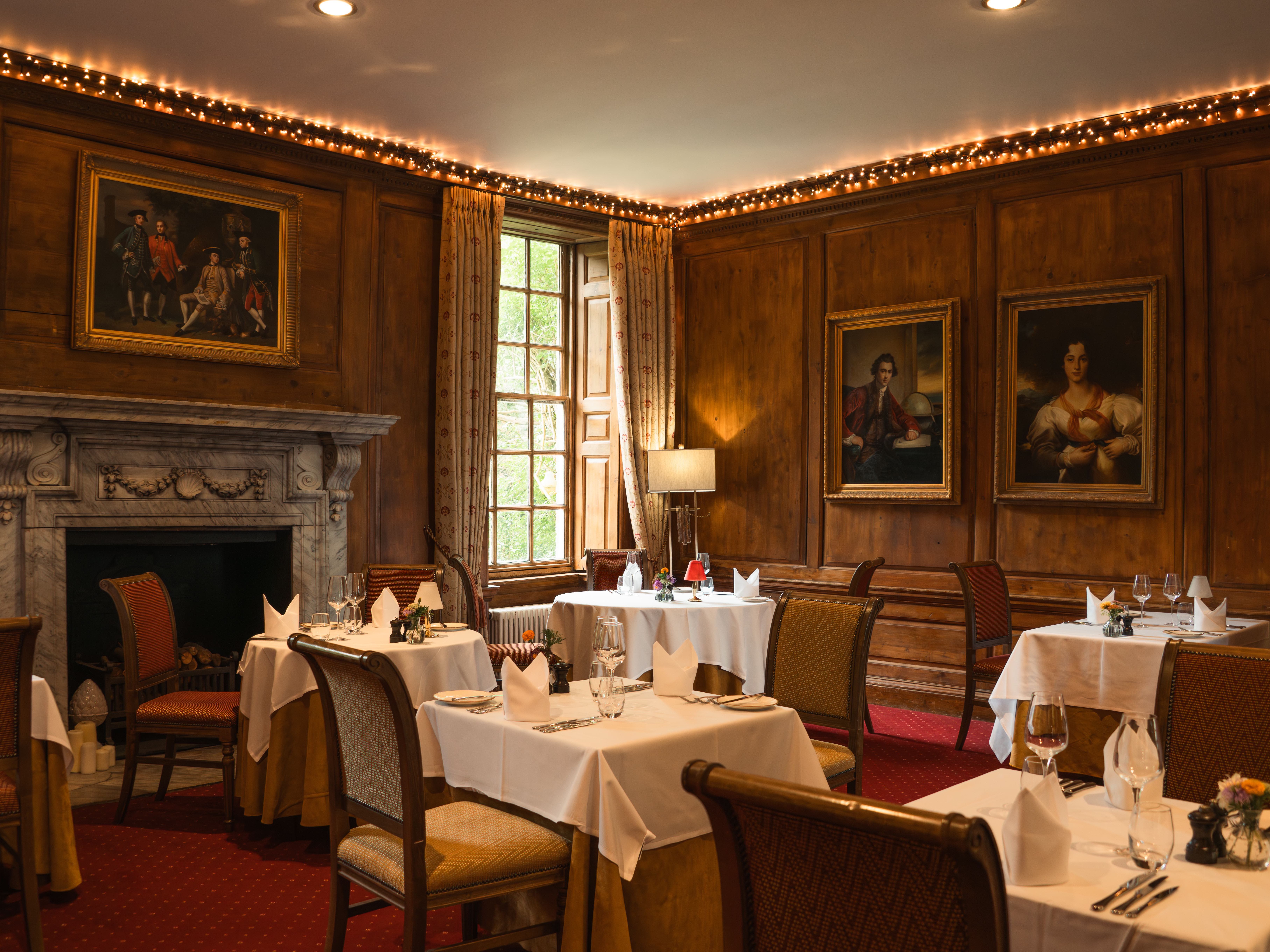 Elegant restaurant dining room with wood-paneled walls, framed portraits, a marble fireplace, and tables set with white tablecloths.