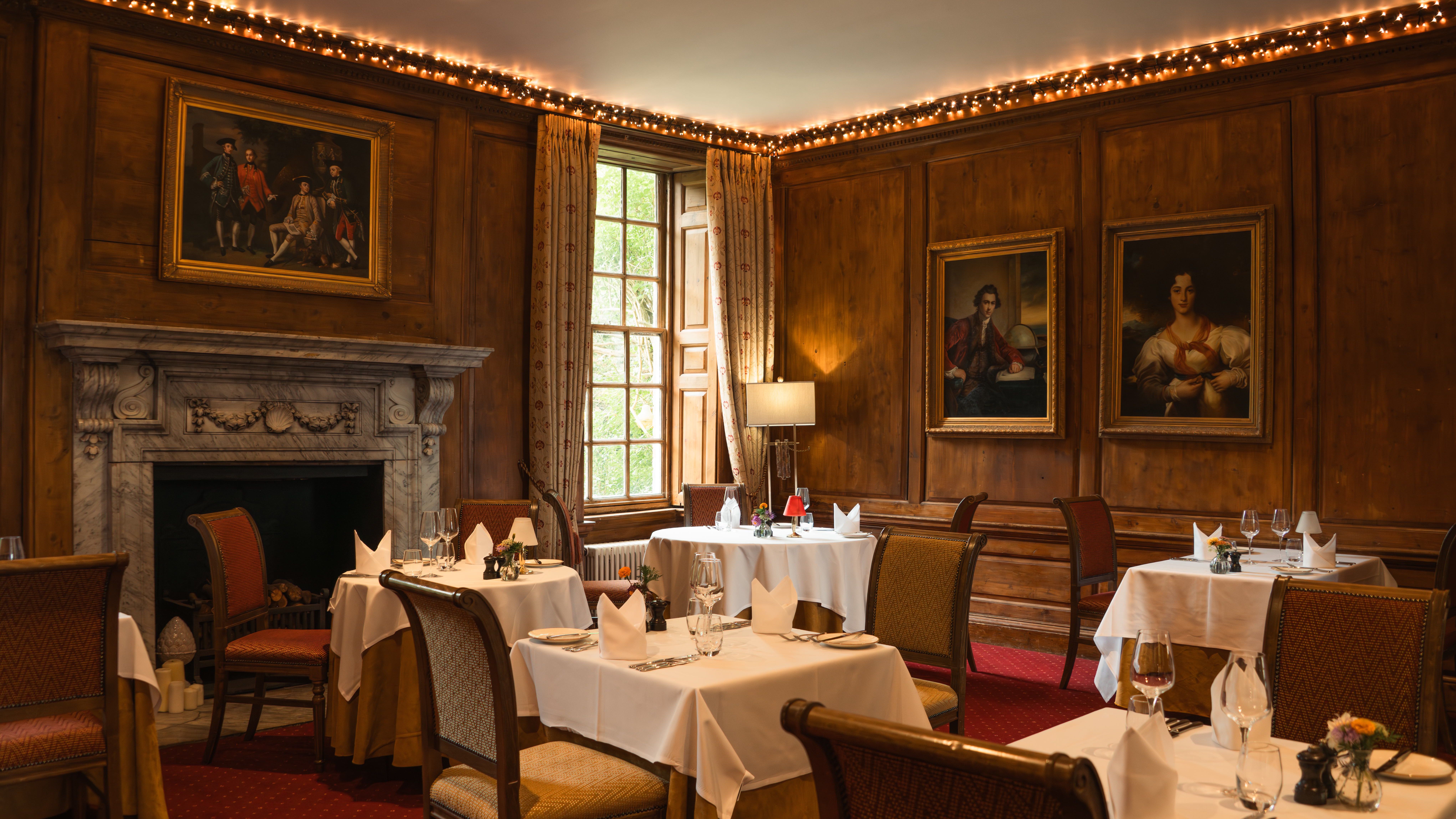 Elegant restaurant dining room with wood-paneled walls, framed portraits, a marble fireplace, and tables set with white tablecloths.