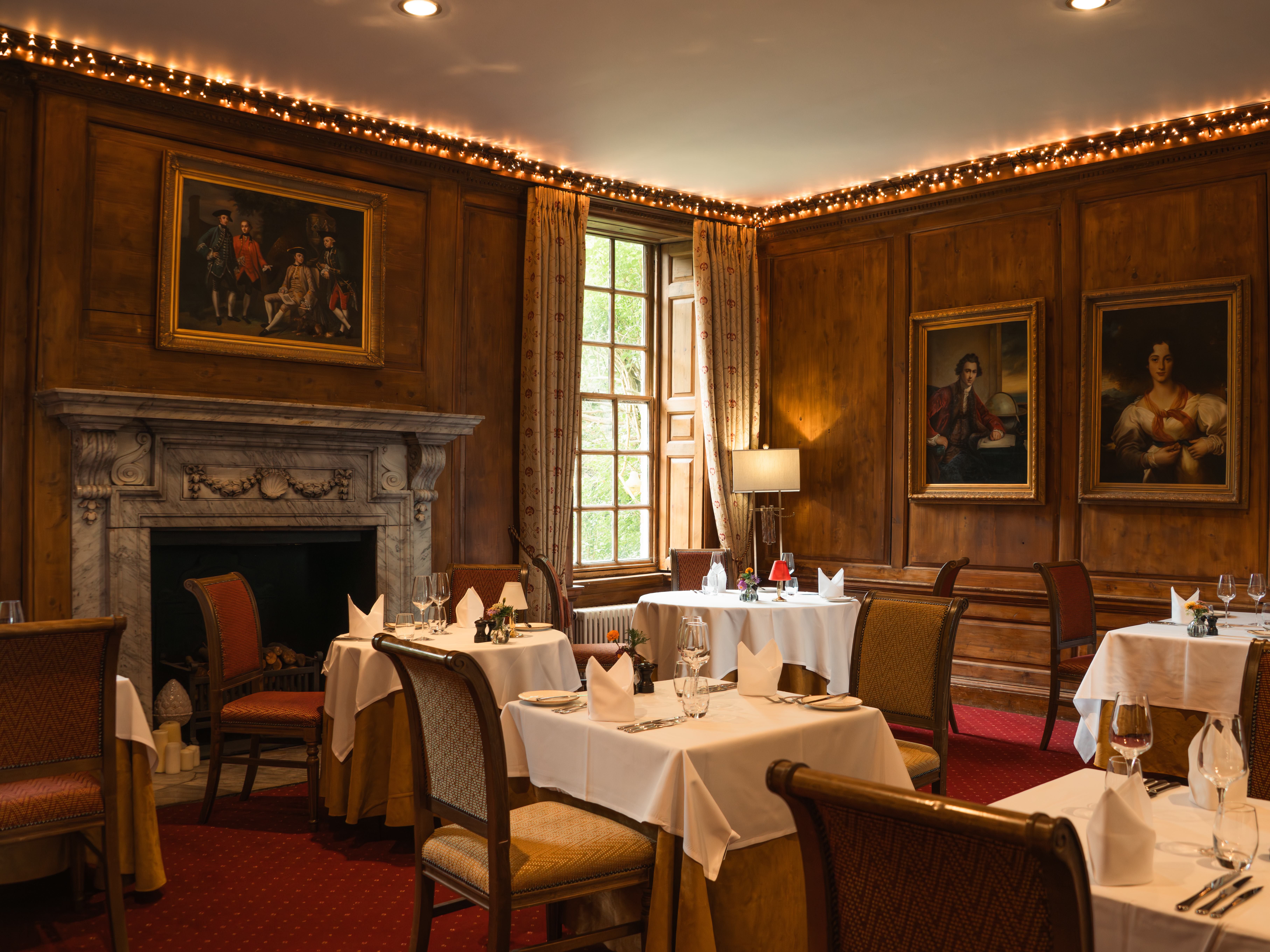 Elegant restaurant dining room with wood-paneled walls, framed portraits, a marble fireplace, and tables set with white tablecloths.