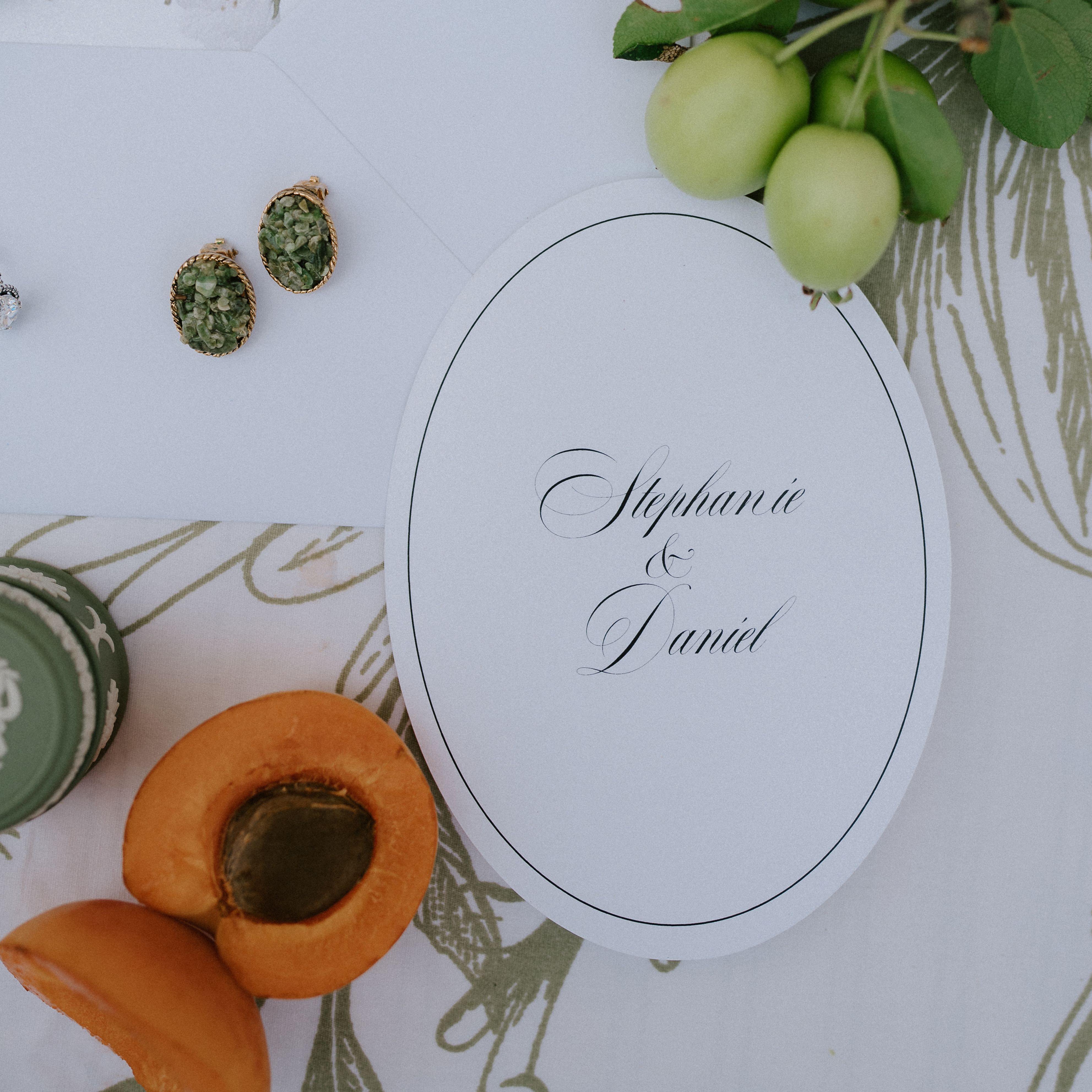 Elegant wedding invitation for Stephanie and Daniel surrounded by apricots, green fruit, floral print, and earrings