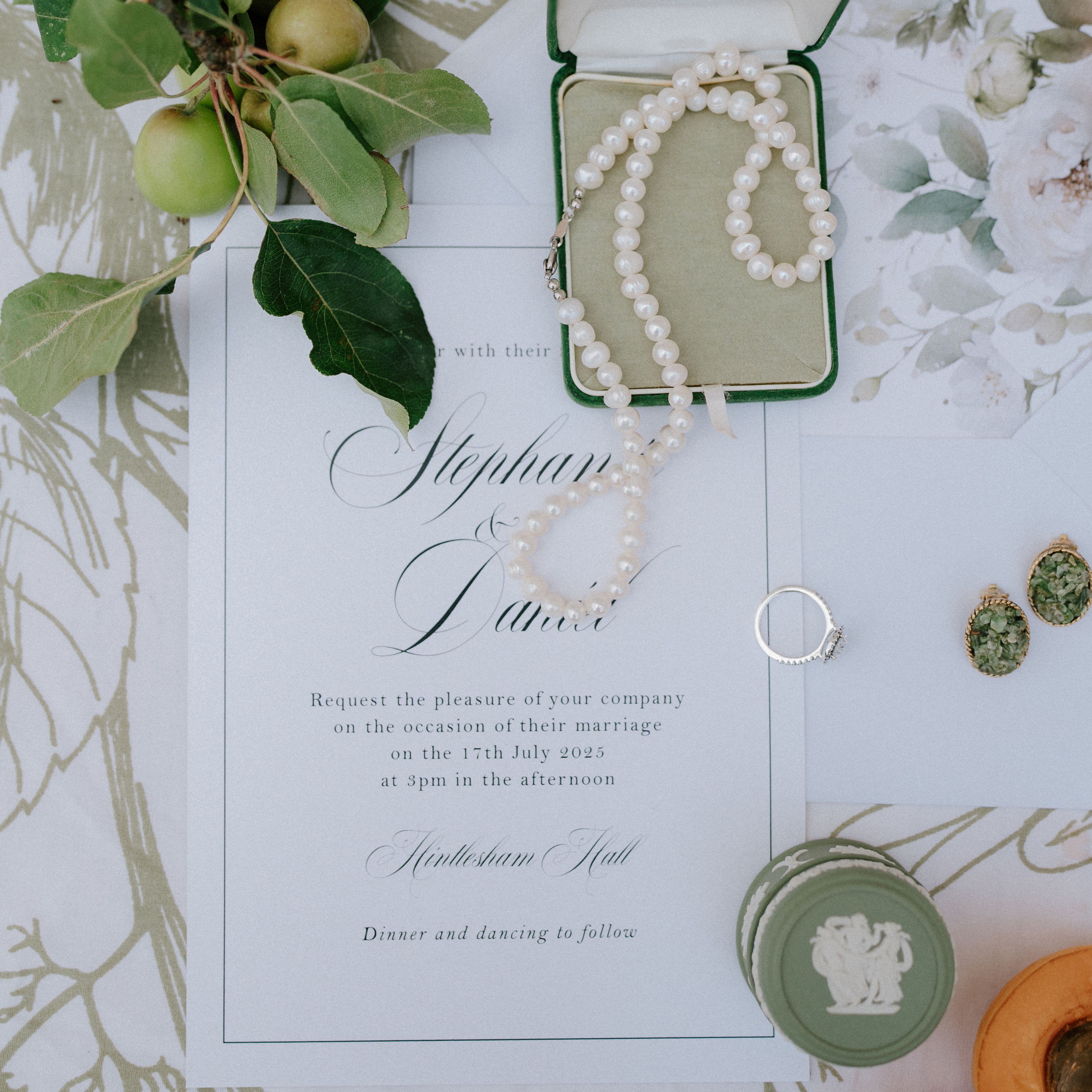 Flat lay of a wedding invitation with pearl necklace, ring, earrings, green apples, and apricots.