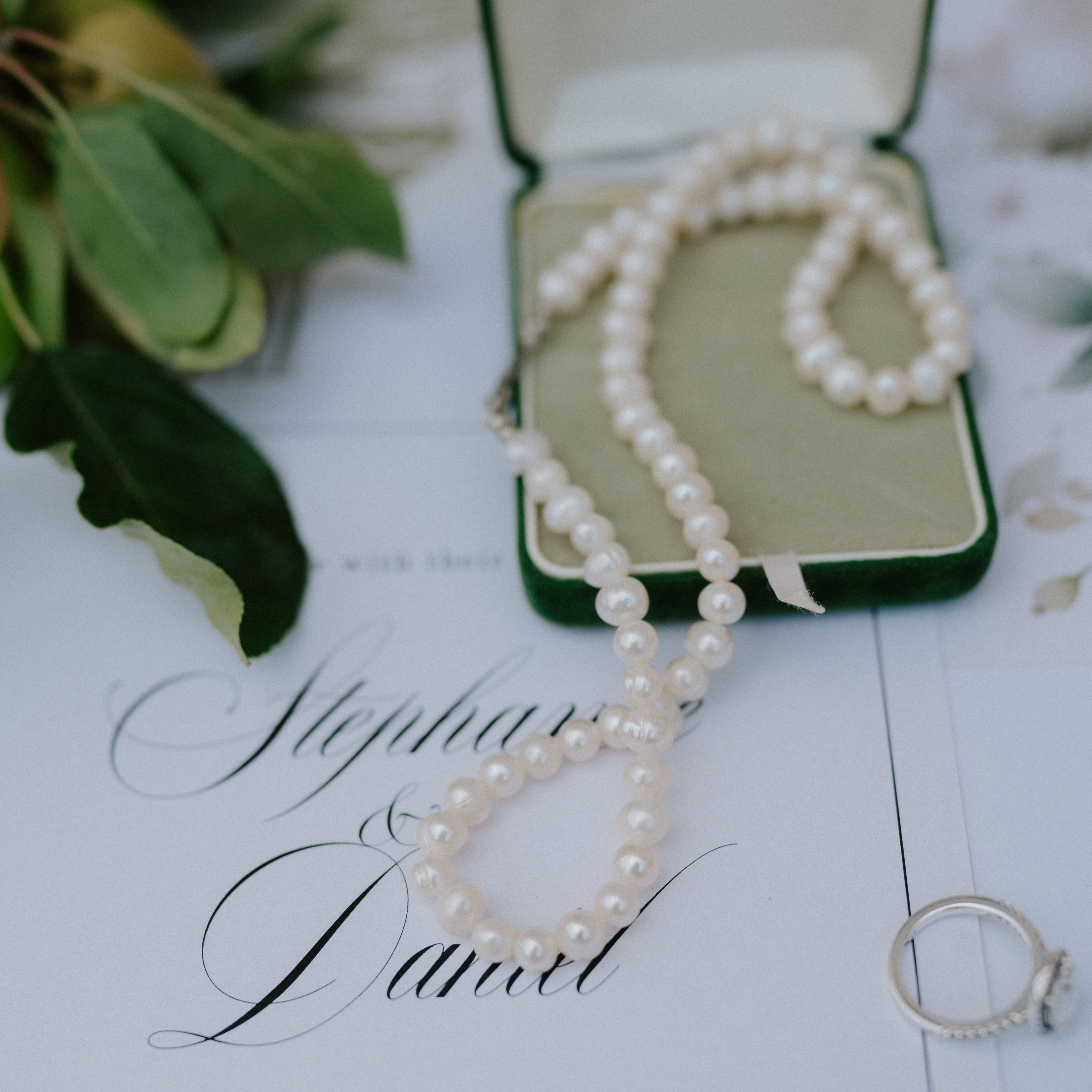 Pearl necklace in a green box, an engagement ring, and a wedding invitation on a table with greenery