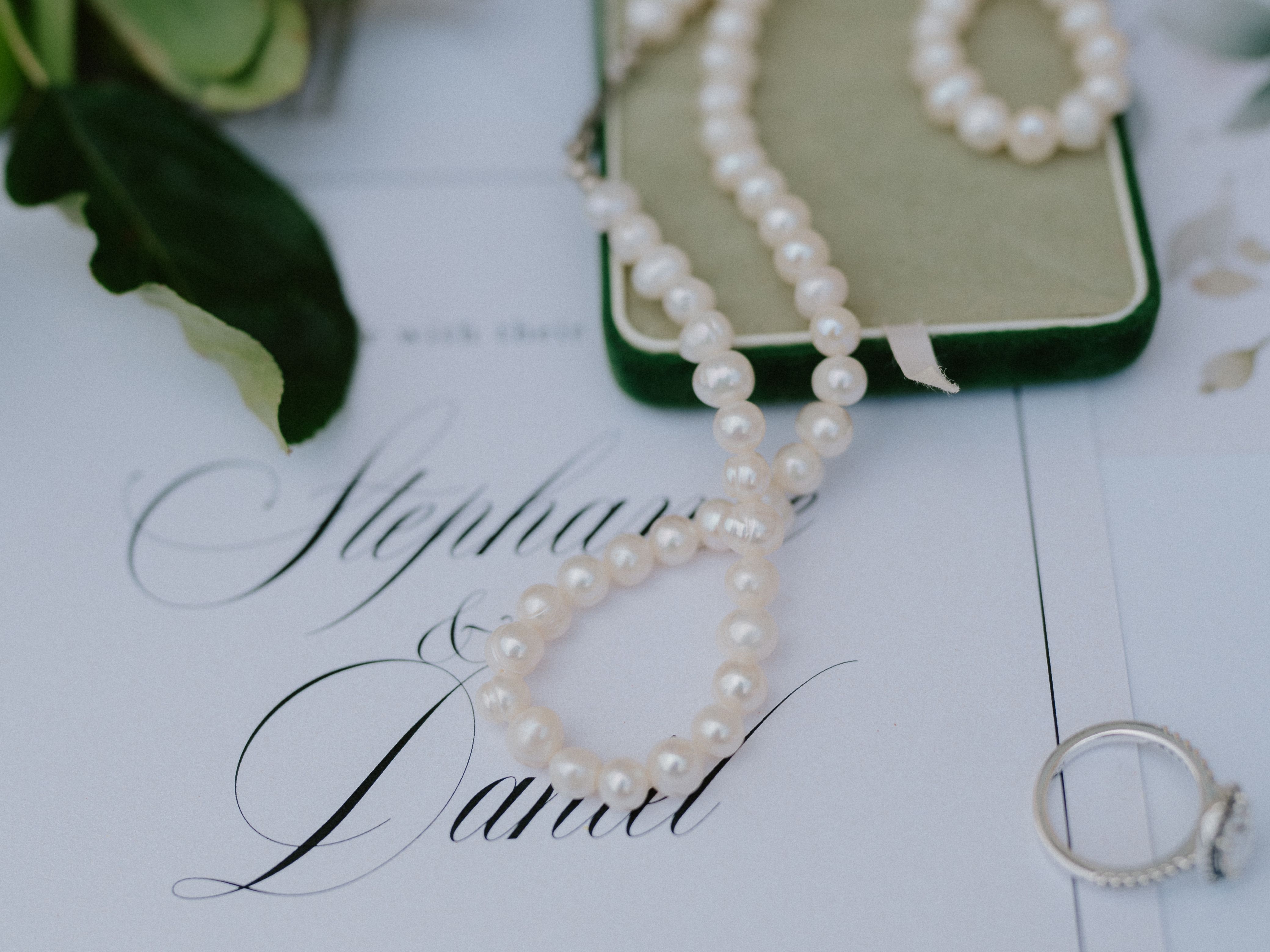 Pearl necklace in a green box, an engagement ring, and a wedding invitation on a table with greenery
