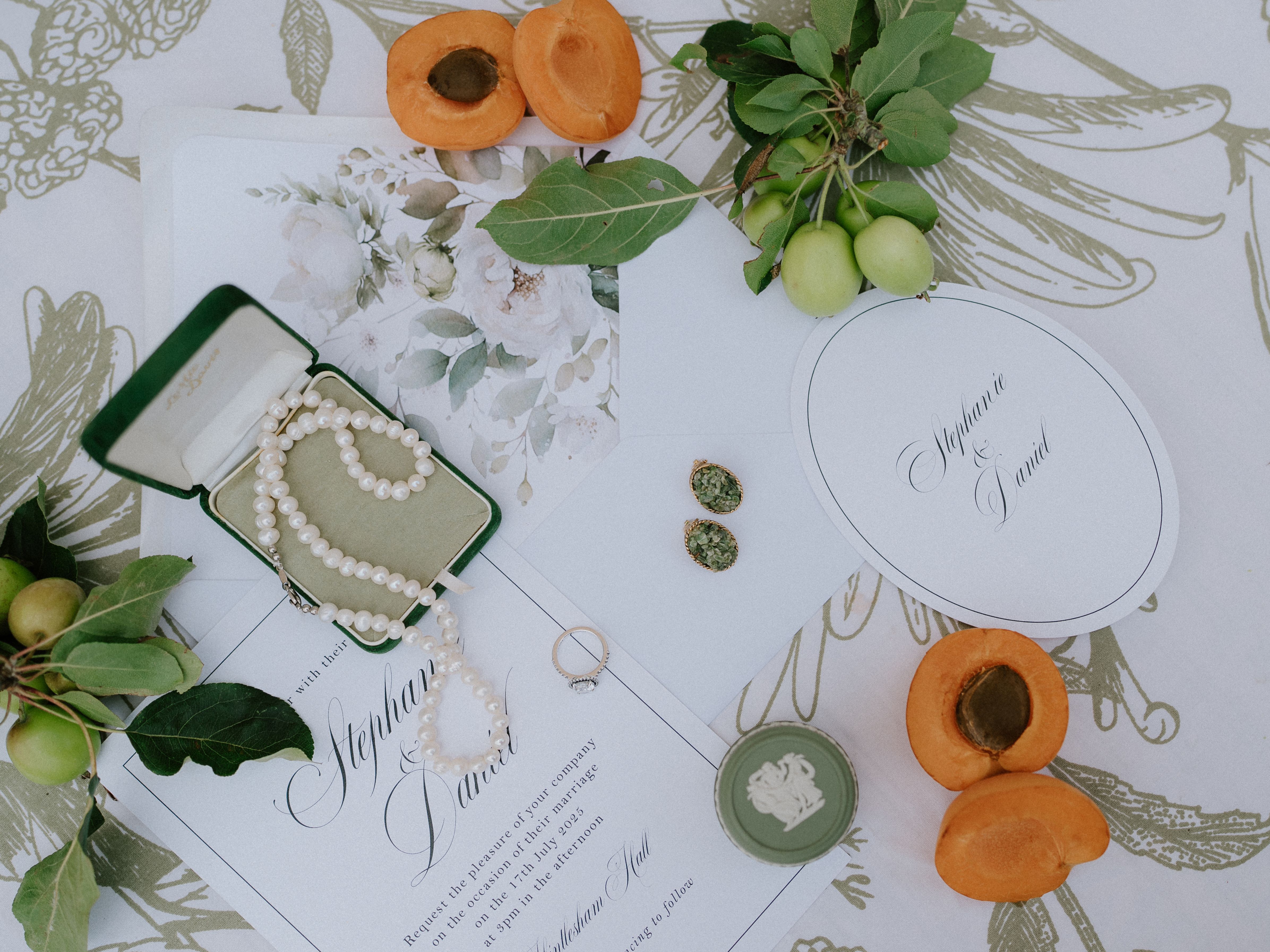 Flat lay of wedding invitations, pearl necklace, apricots, jewelry, and green fruit on a floral tablecloth.