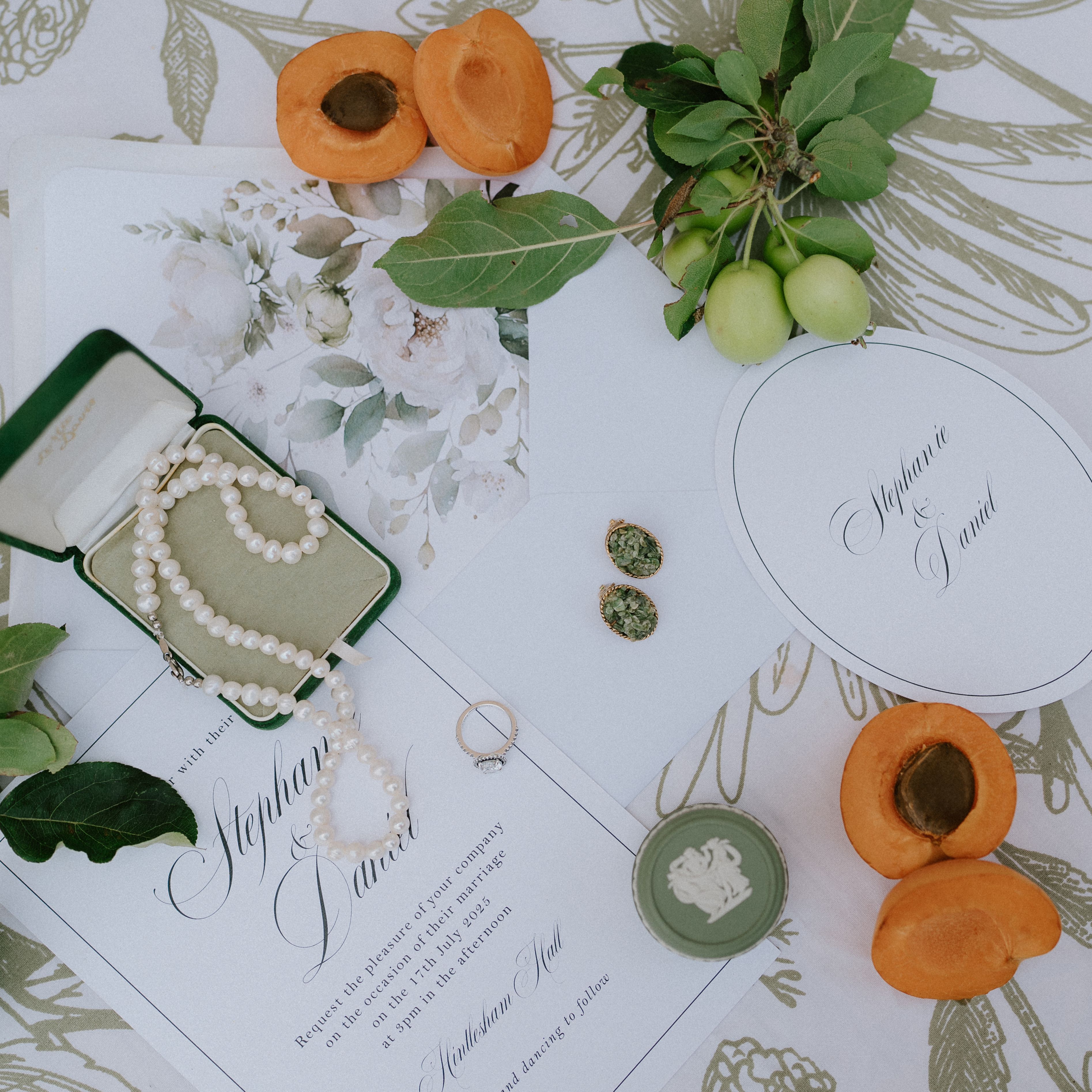 Flat lay of wedding invitations, pearl necklace, apricots, jewelry, and green fruit on a floral tablecloth.