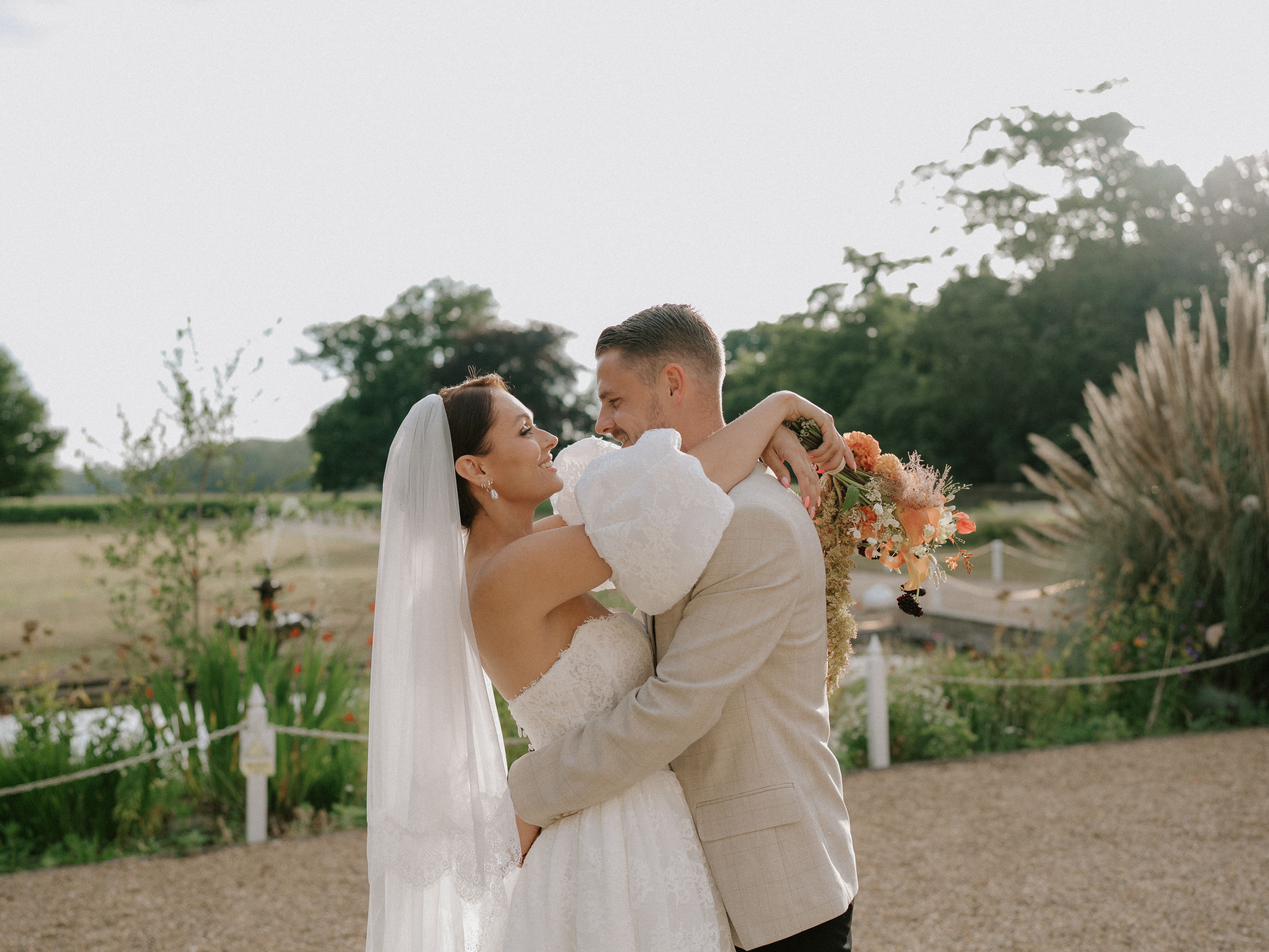 Bride and groom embracing outdoors on their wedding day, with the bride holding a bouquet of flowers.