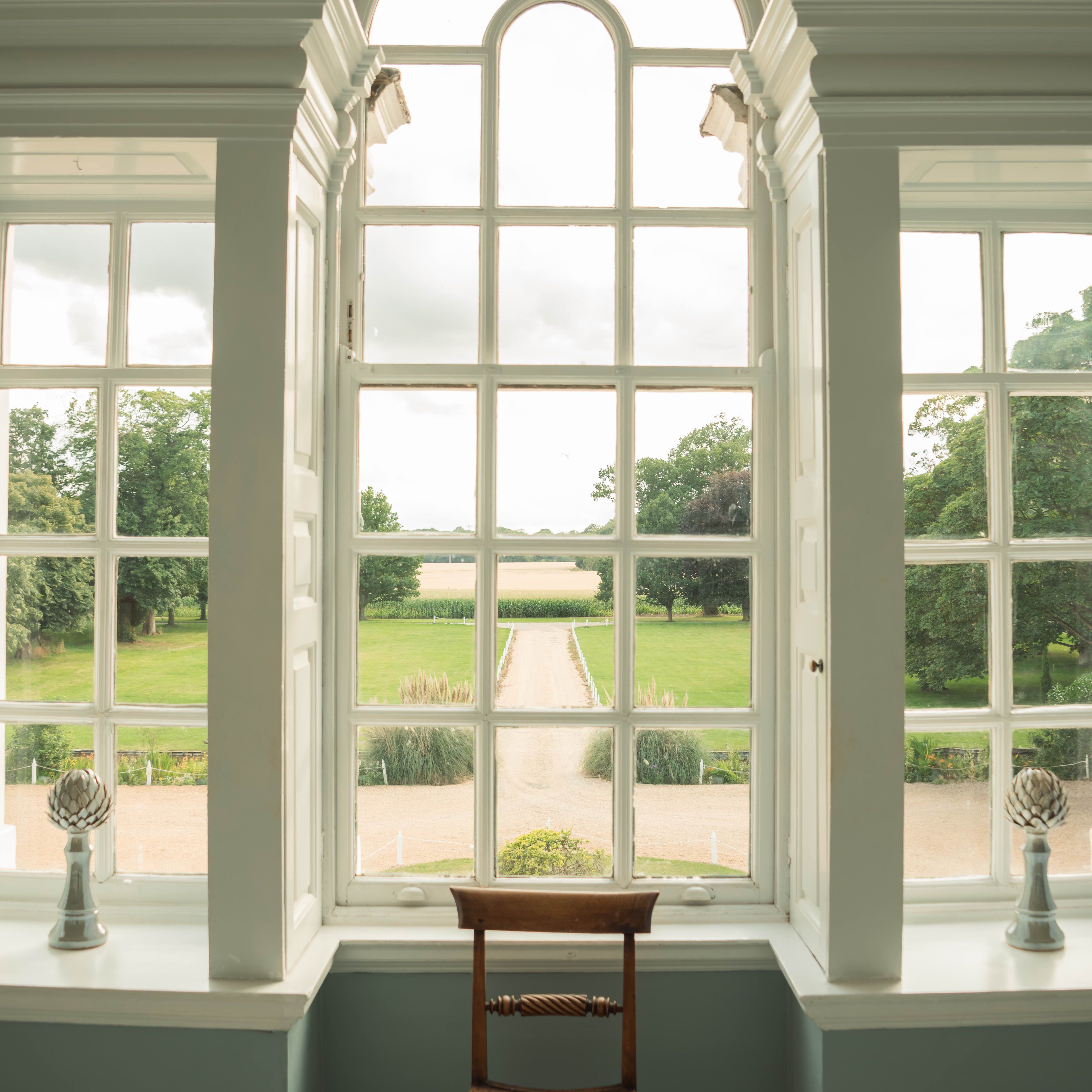 A wooden chair placed in front of a large arched window with a view of a garden and gravel path outside.