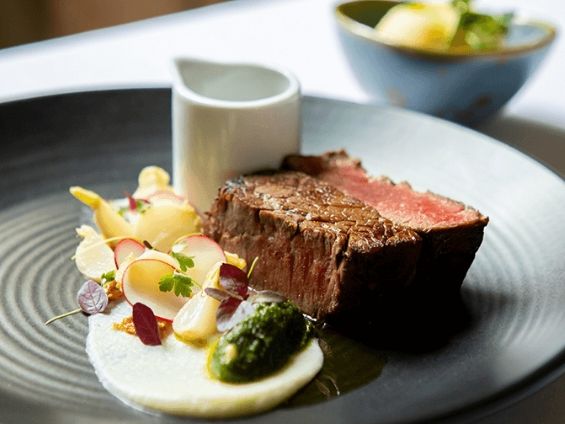 Gourmet plated dish featuring a medium-rare steak with vegetable garnishes and sauce on a black plate