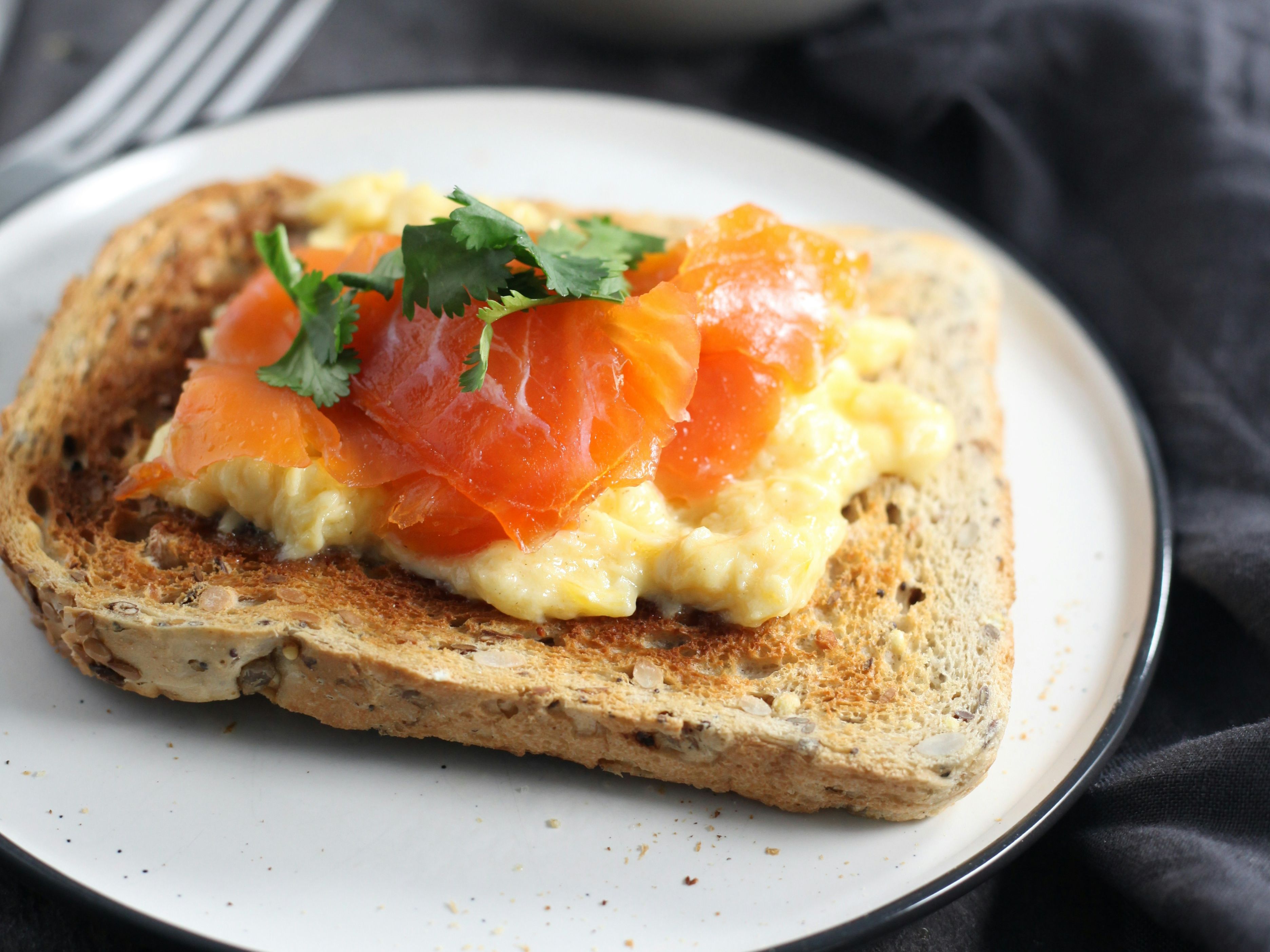Scrambled eggs and smoked salmon on multigrain toast, garnished with fresh herbs, served on a white plate.