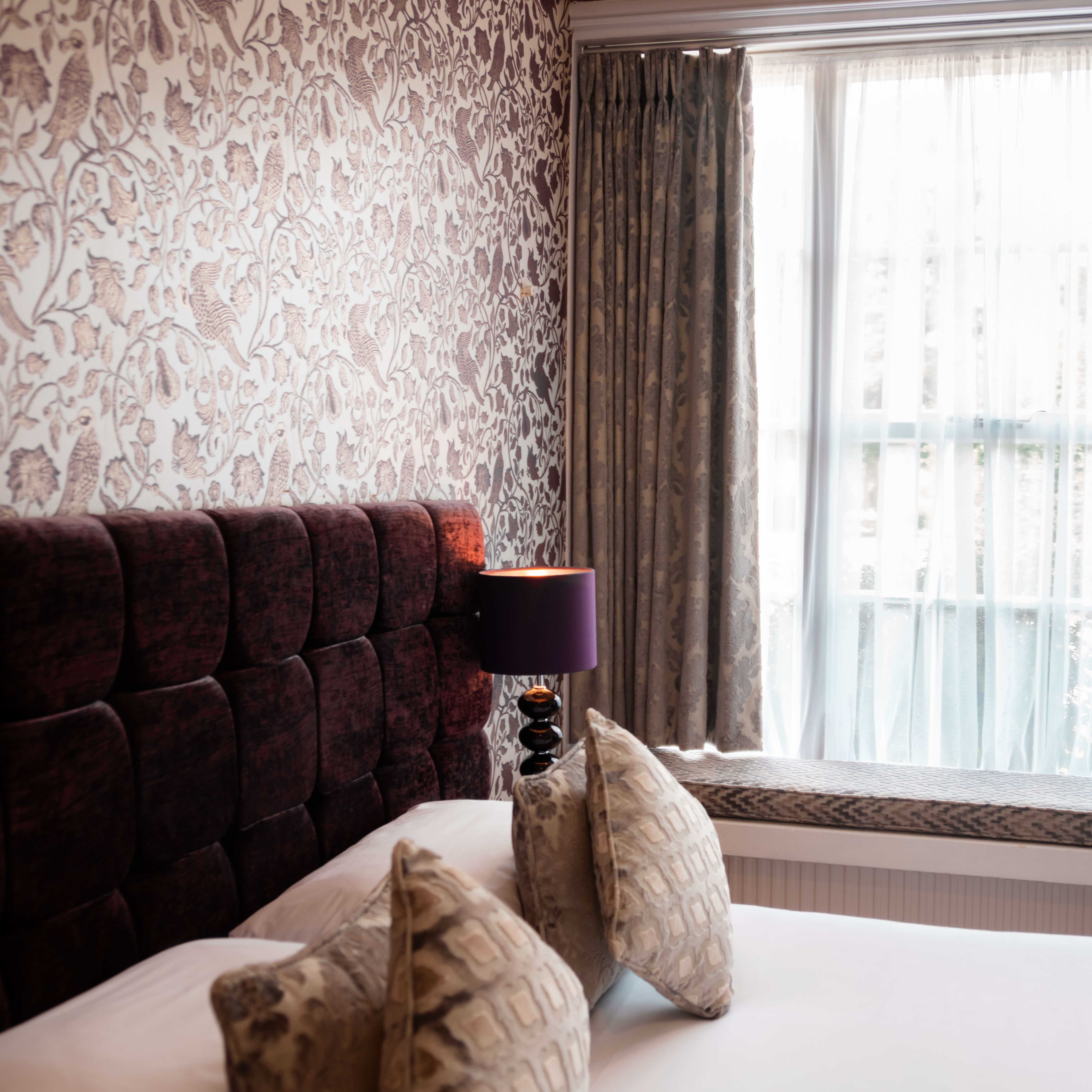 Cozy bedroom with patterned wallpaper, upholstered headboard, and window with sheer and heavy curtains.