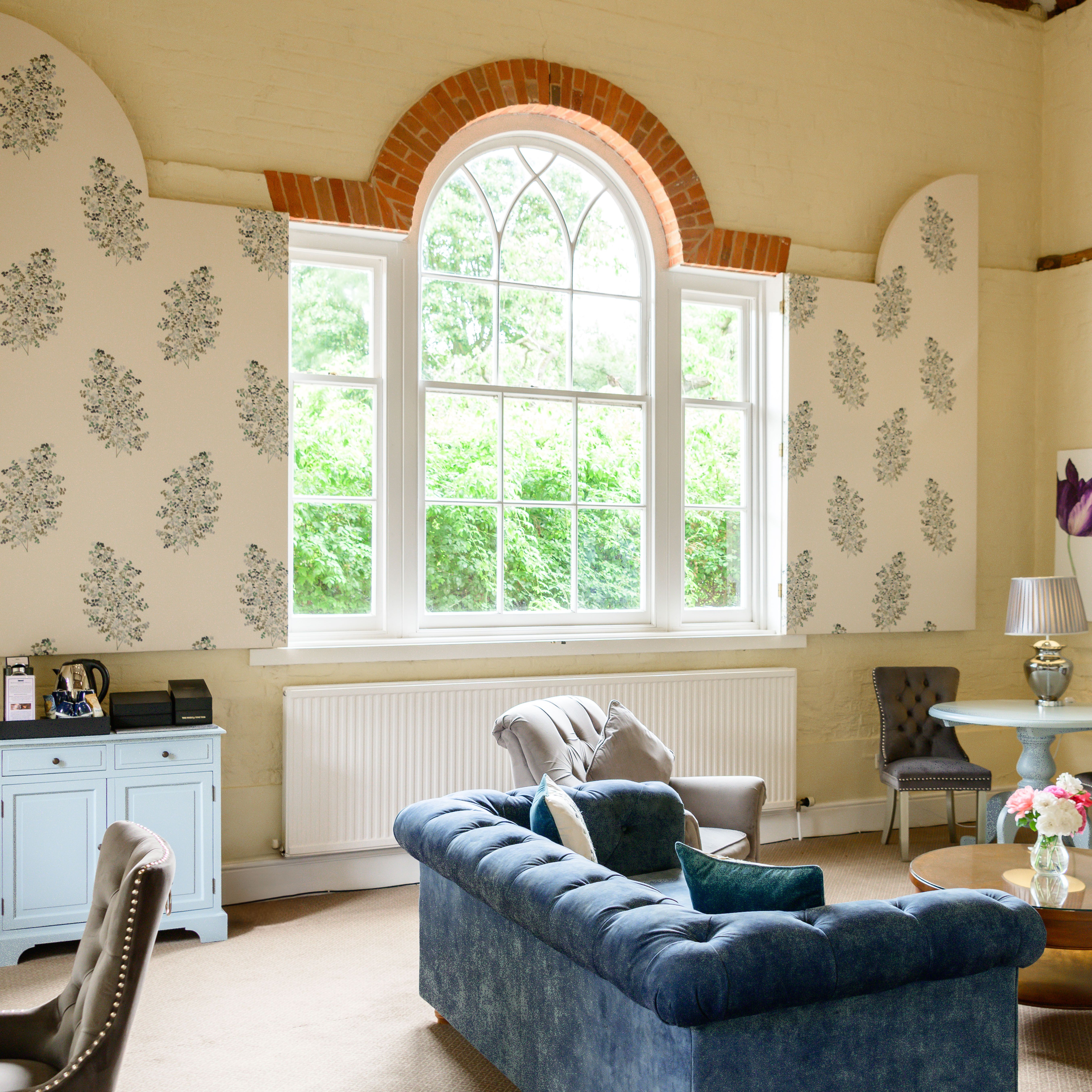 Brightly lit living room with large arched window, blue velvet sofa, and floral decor.