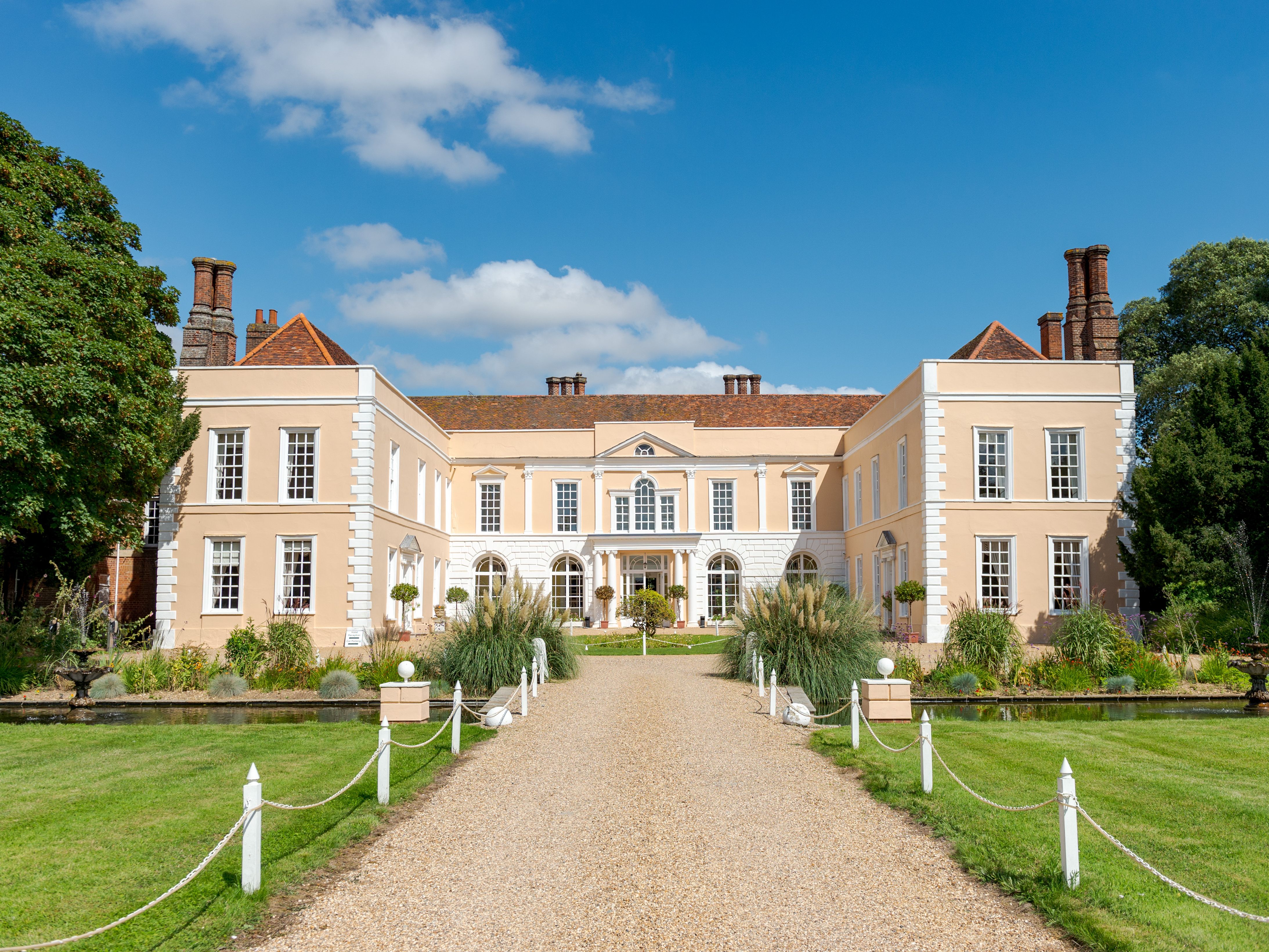 Large elegant mansion with beige walls, white trim, and red roof, surrounded by manicured gardens, fountains, and a gravel path under blue sky