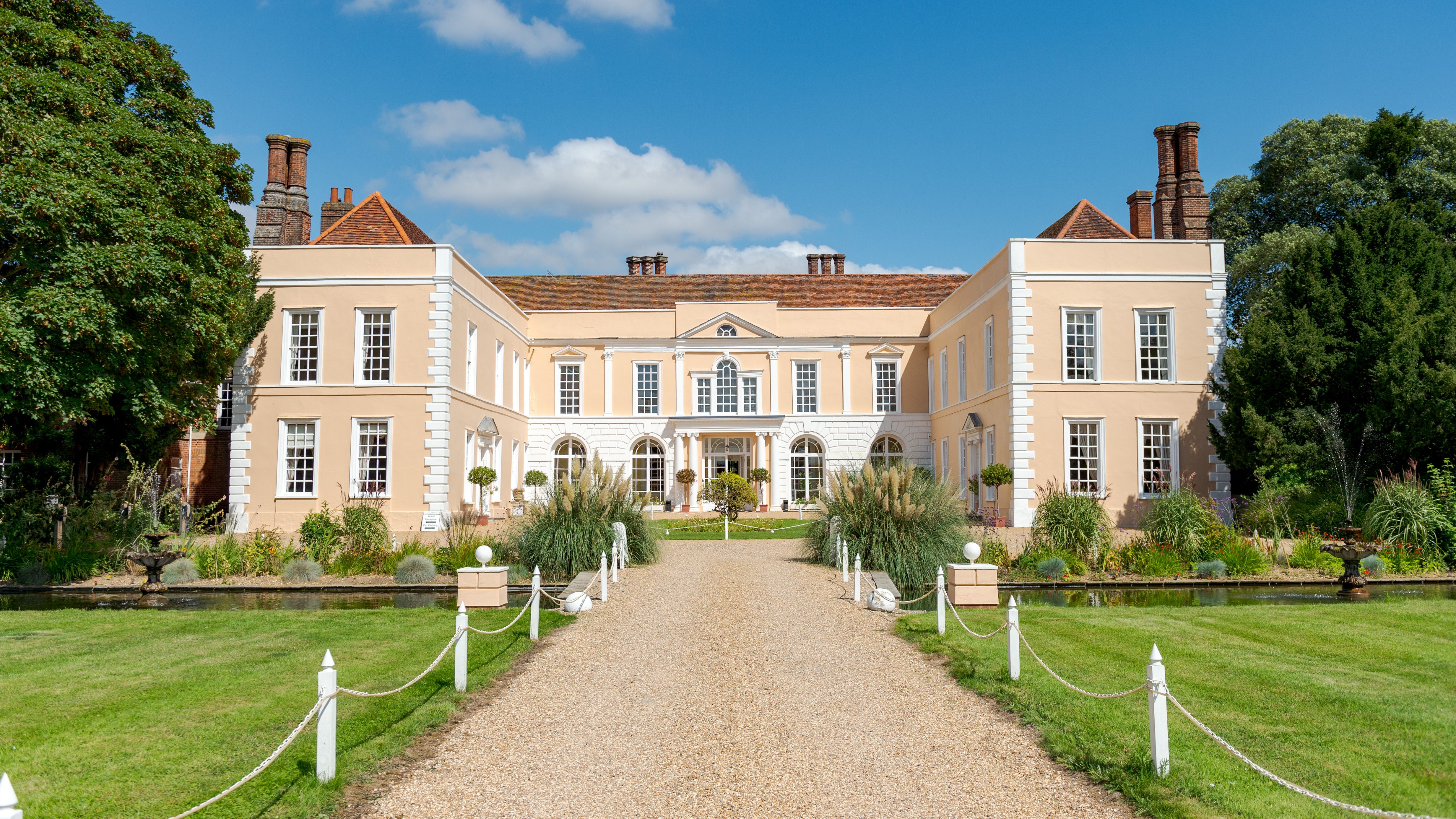 Large elegant mansion with beige walls, white trim, and red roof, surrounded by manicured gardens, fountains, and a gravel path under blue sky