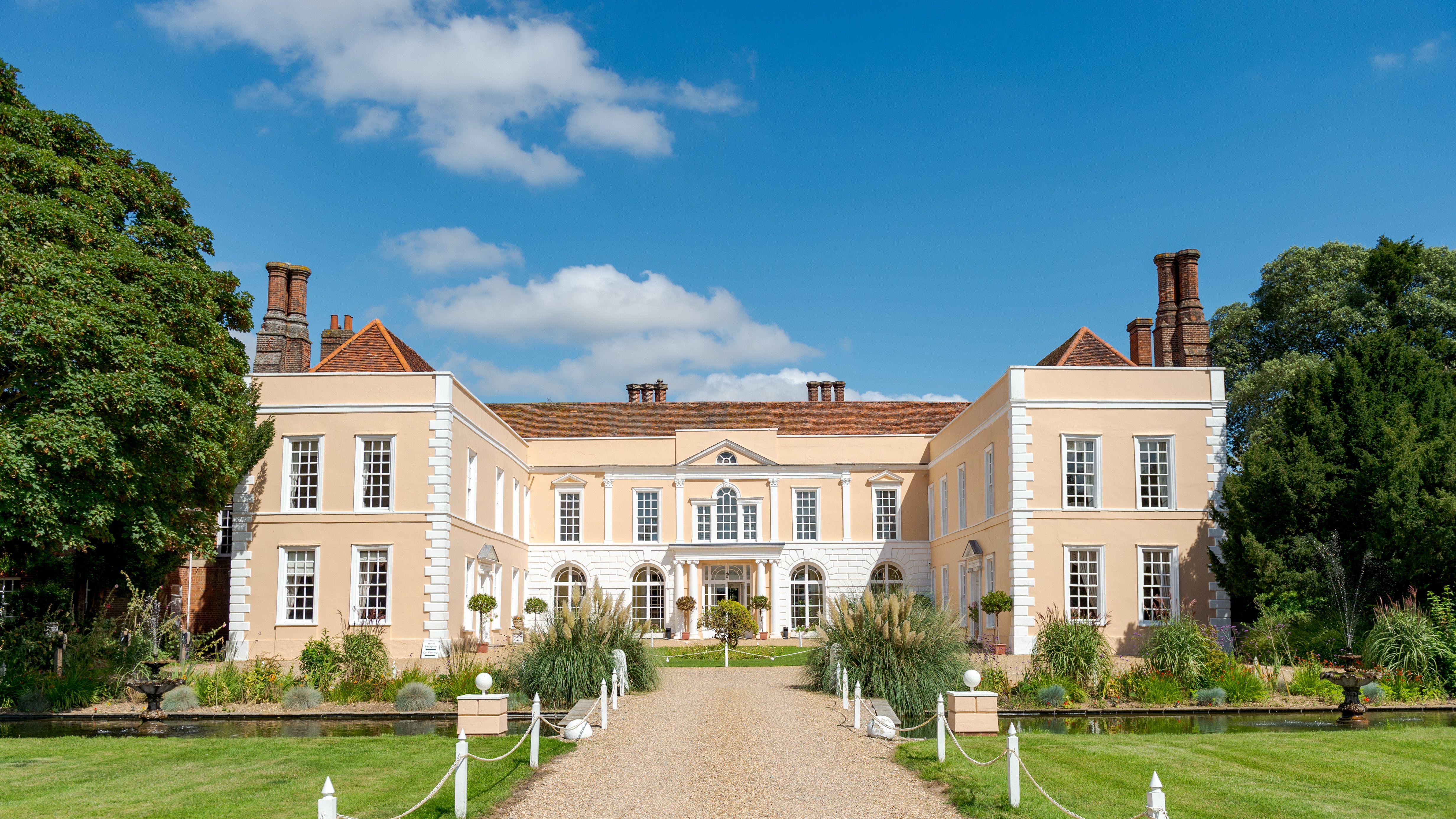 Large elegant mansion with beige walls, white trim, and red roof, surrounded by manicured gardens, fountains, and a gravel path under blue sky