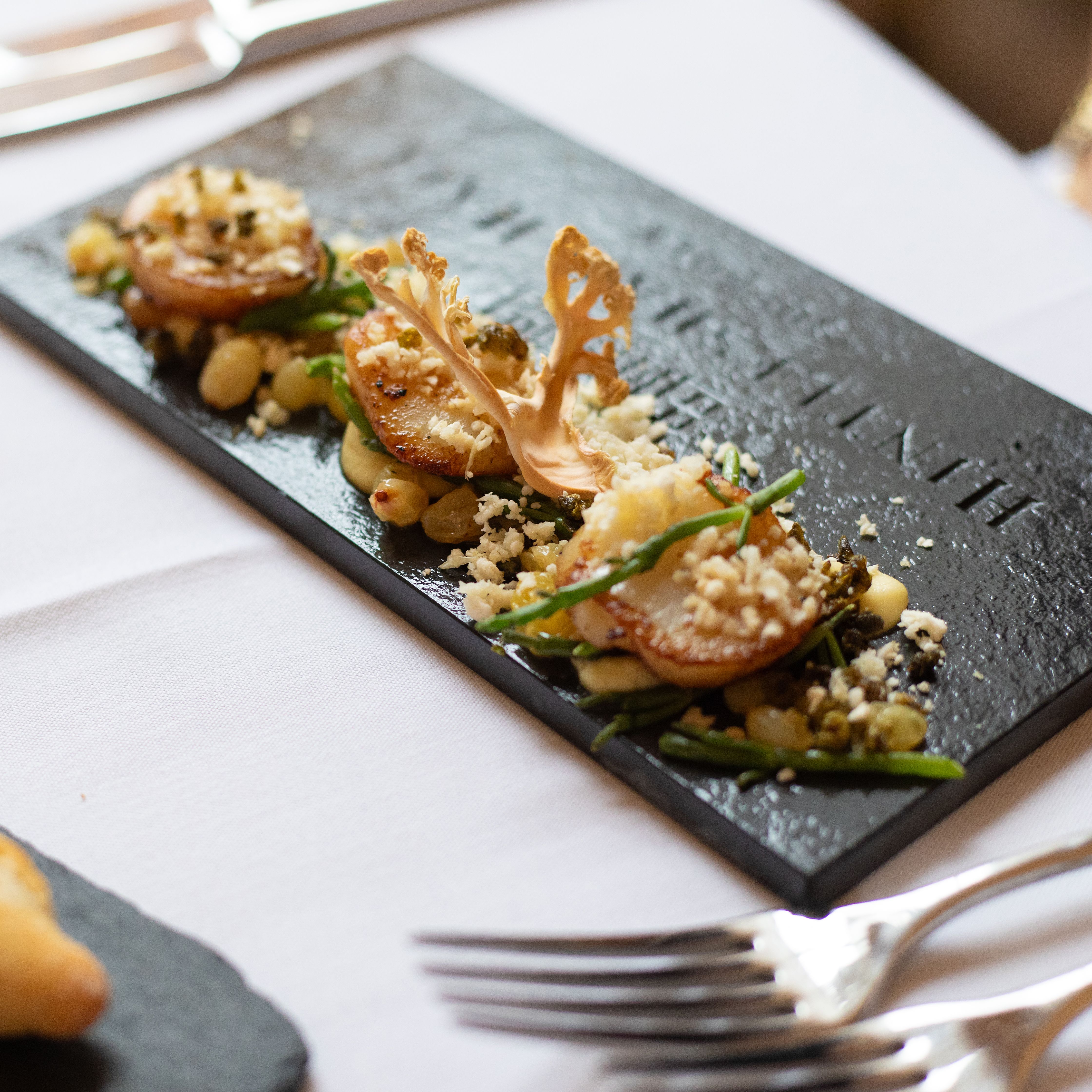 A gourmet dish artfully plated on a rectangular black slate, with roasted vegetables, crumbs, a decorative crisp, and herbs. A bread roll and cutlery are also visible on a white tablecloth.