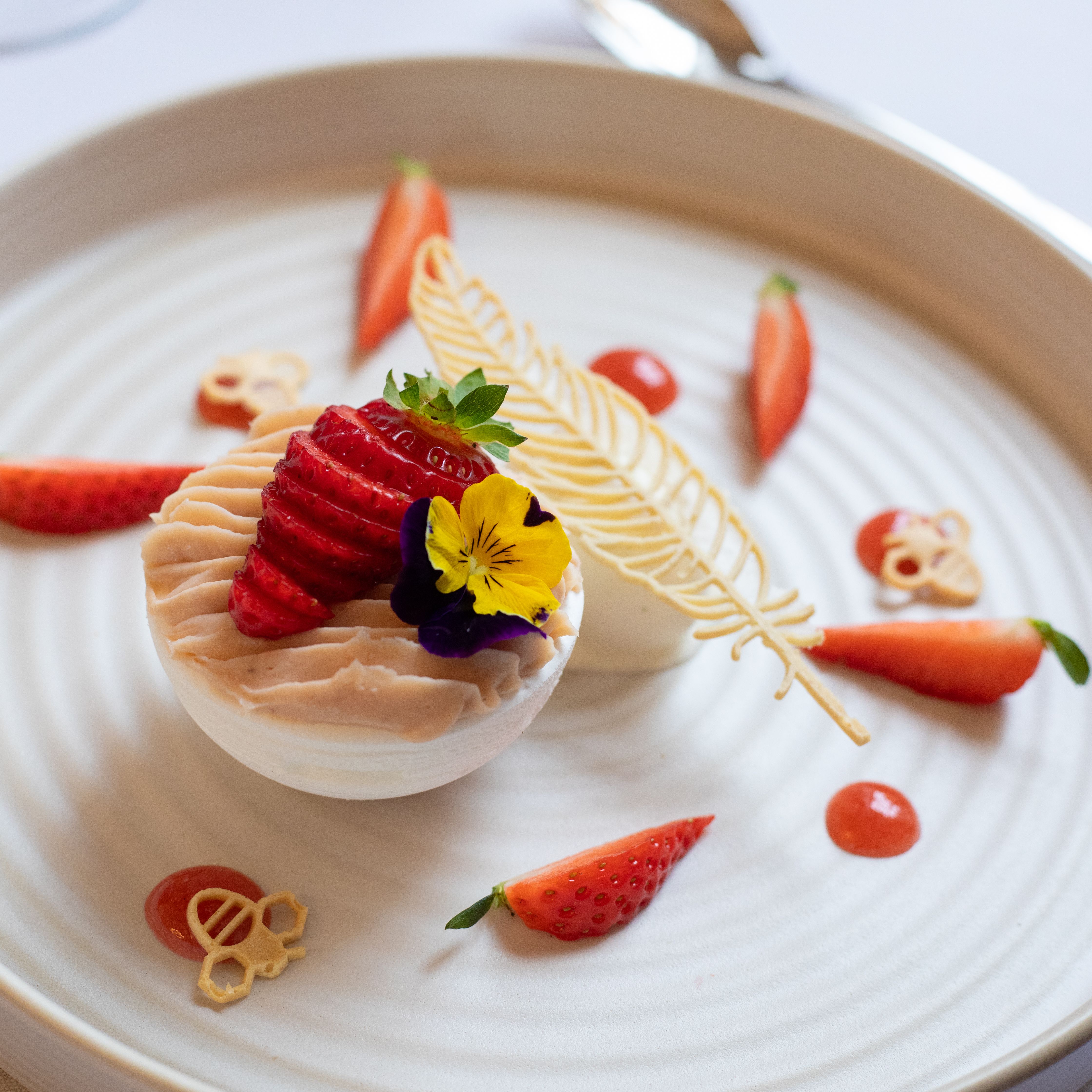 Elegant plated dessert with decorative elements, fresh strawberries, and edible flowers on a round white plate.