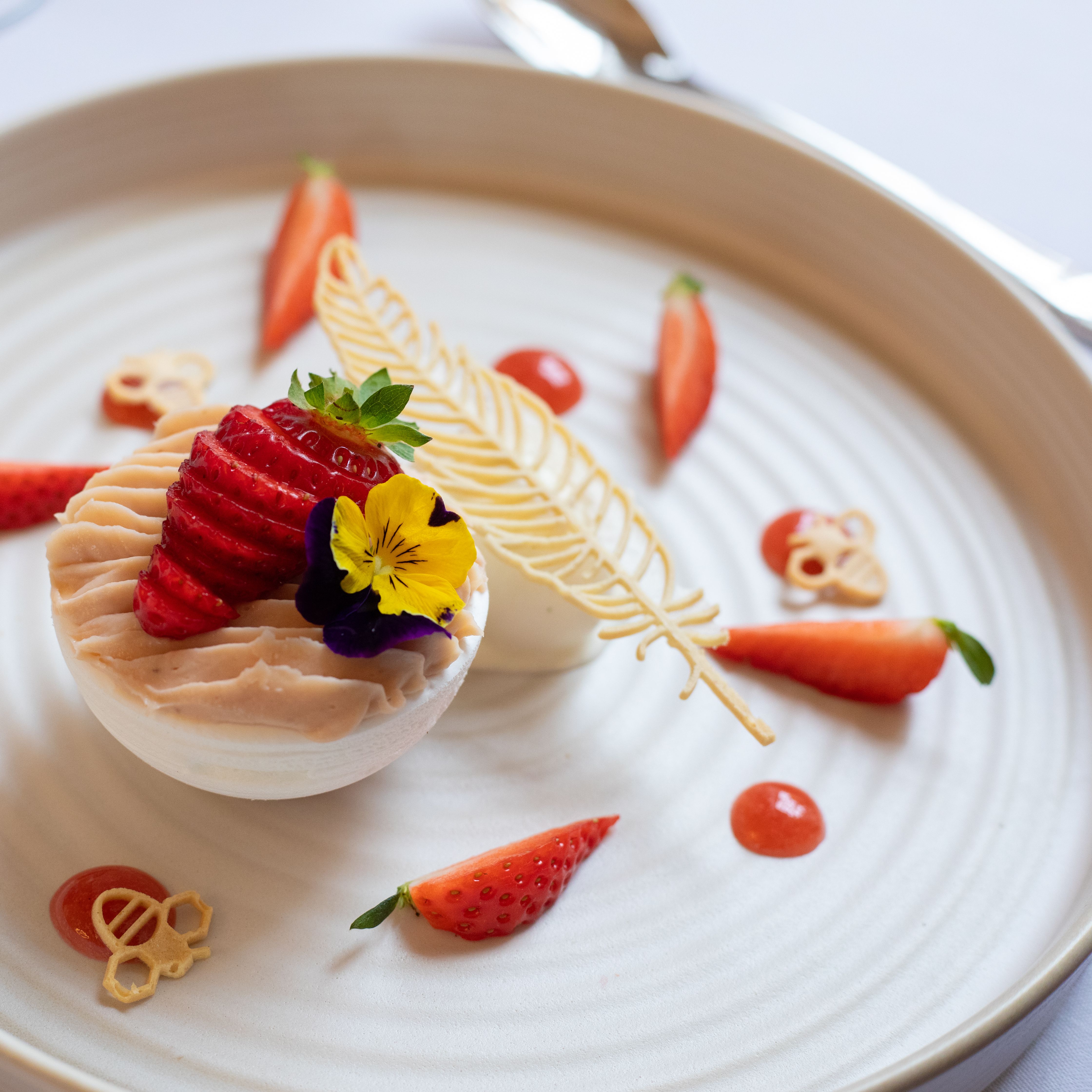 Elegant plated dessert with decorative elements, fresh strawberries, and edible flowers on a round white plate.