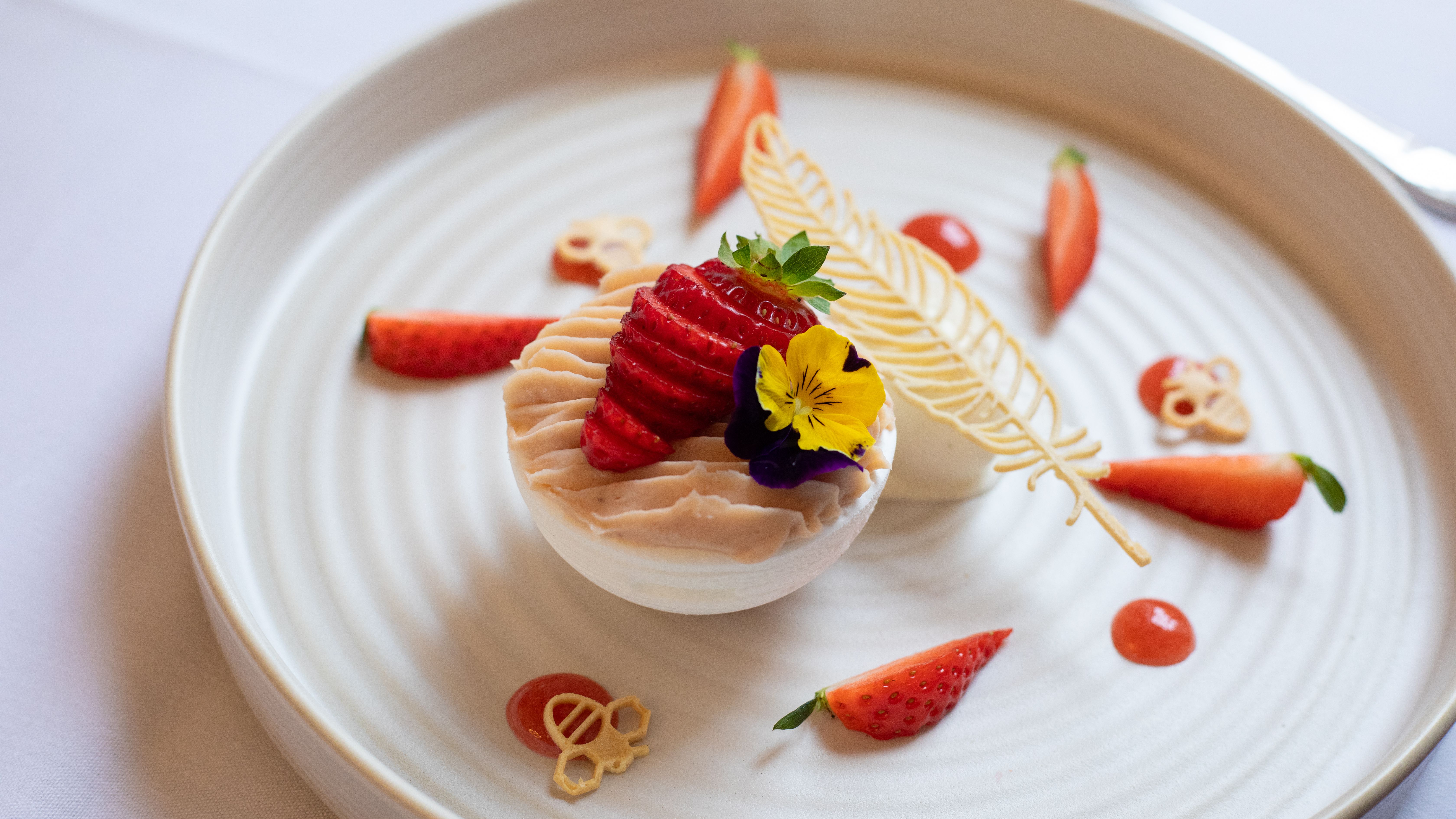 Elegant plated dessert with decorative elements, fresh strawberries, and edible flowers on a round white plate.