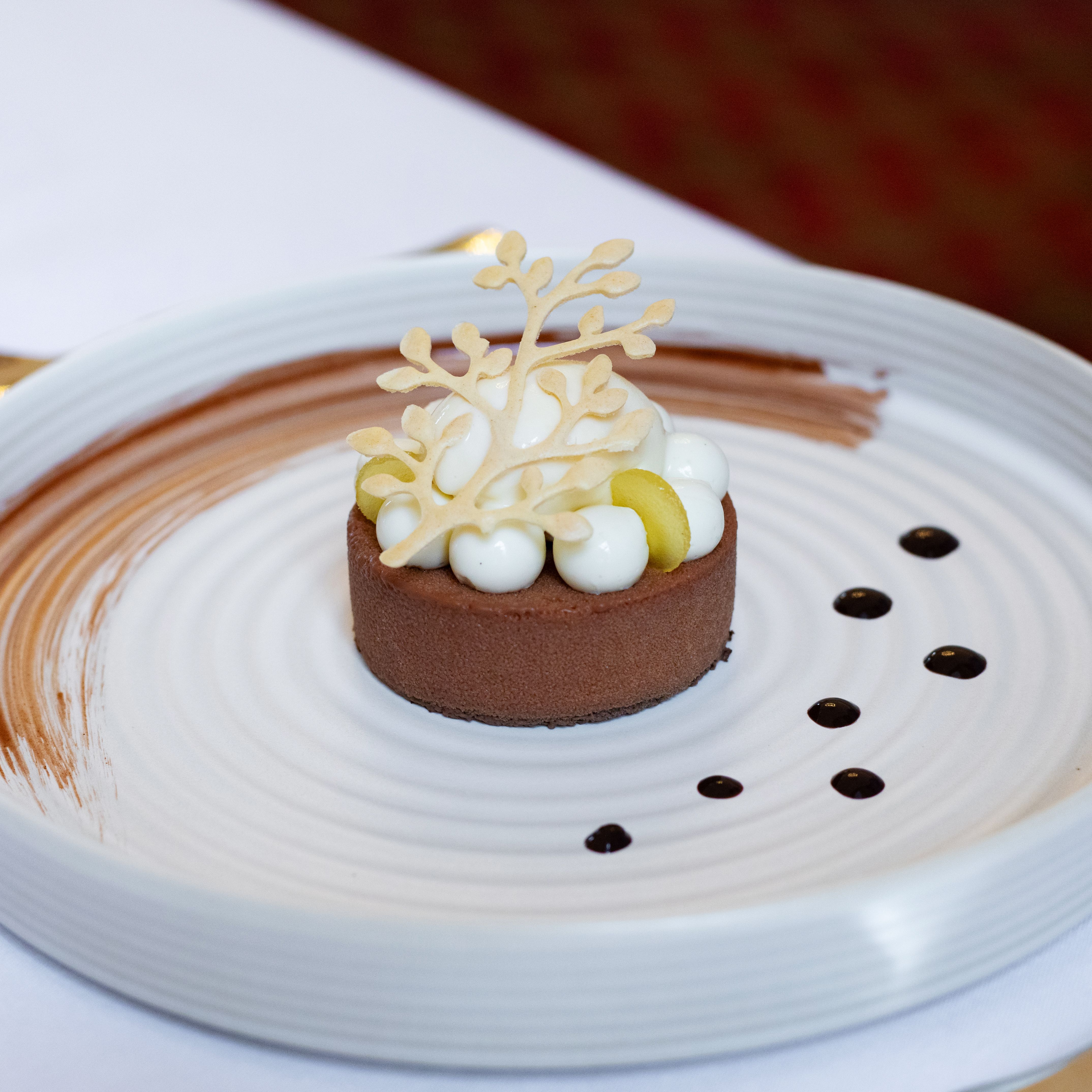 A beautifully plated chocolate dessert garnished with white cream, yellow spheres, and a decorative white chocolate topper.