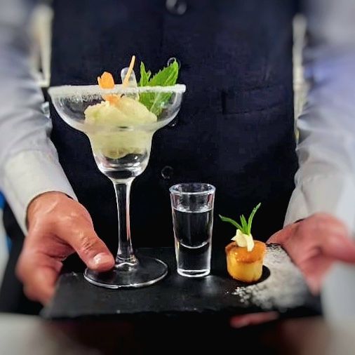 A server presenting a gourmet dessert on a slate tray featuring a cocktail glass with a sorbet, a shot glass with a clear beverage, and a small pastry with garnish.