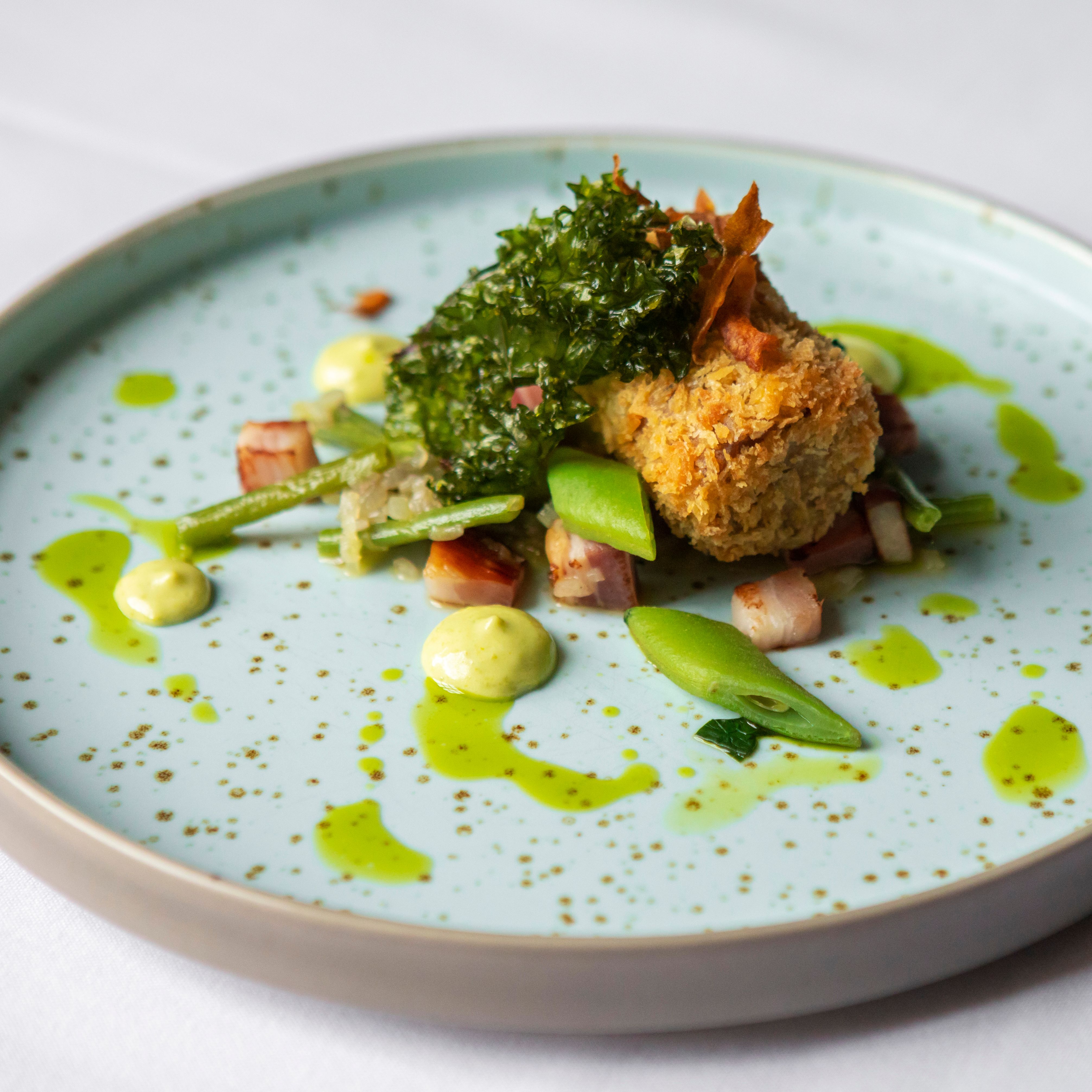 Plated gourmet dish featuring a golden-brown croquette with vegetables and green sauce drizzled on a speckled plate