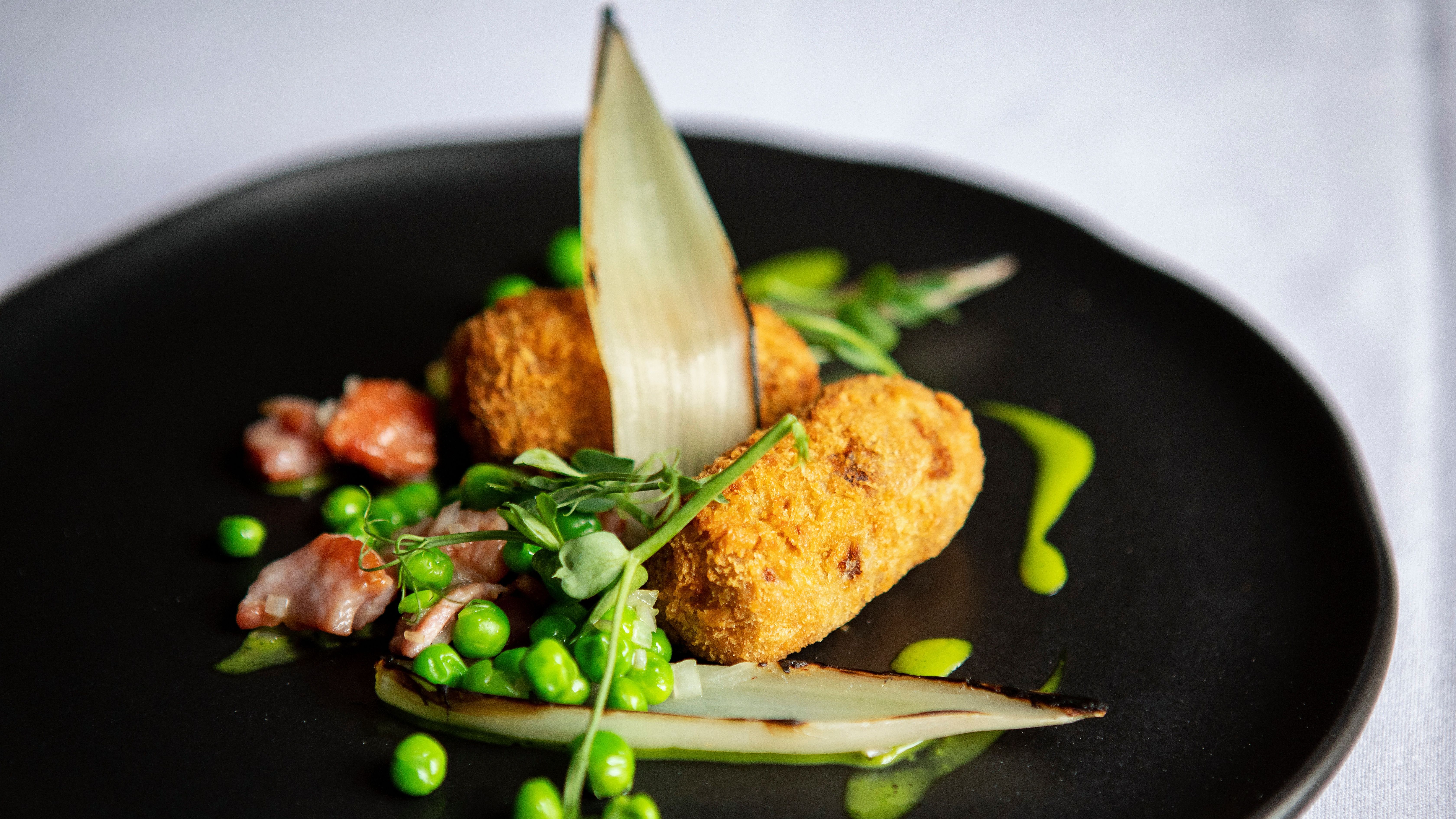 Gourmet dish with croquettes, peas, greens, and bacon on a black plate