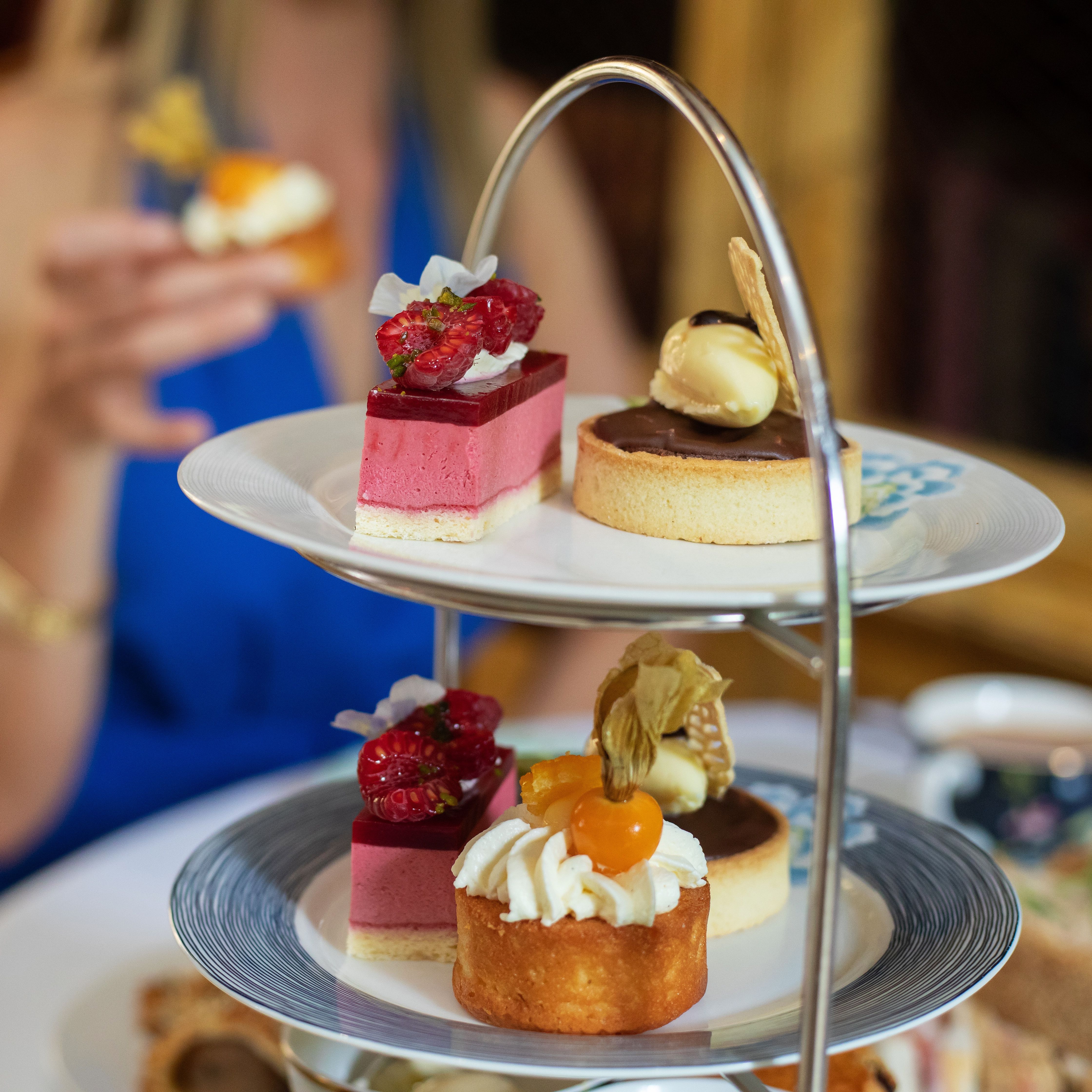 Afternoon tea table with a woman in a blue dress holding a small cake, and a tiered stand filled with colorful desserts and pastries.