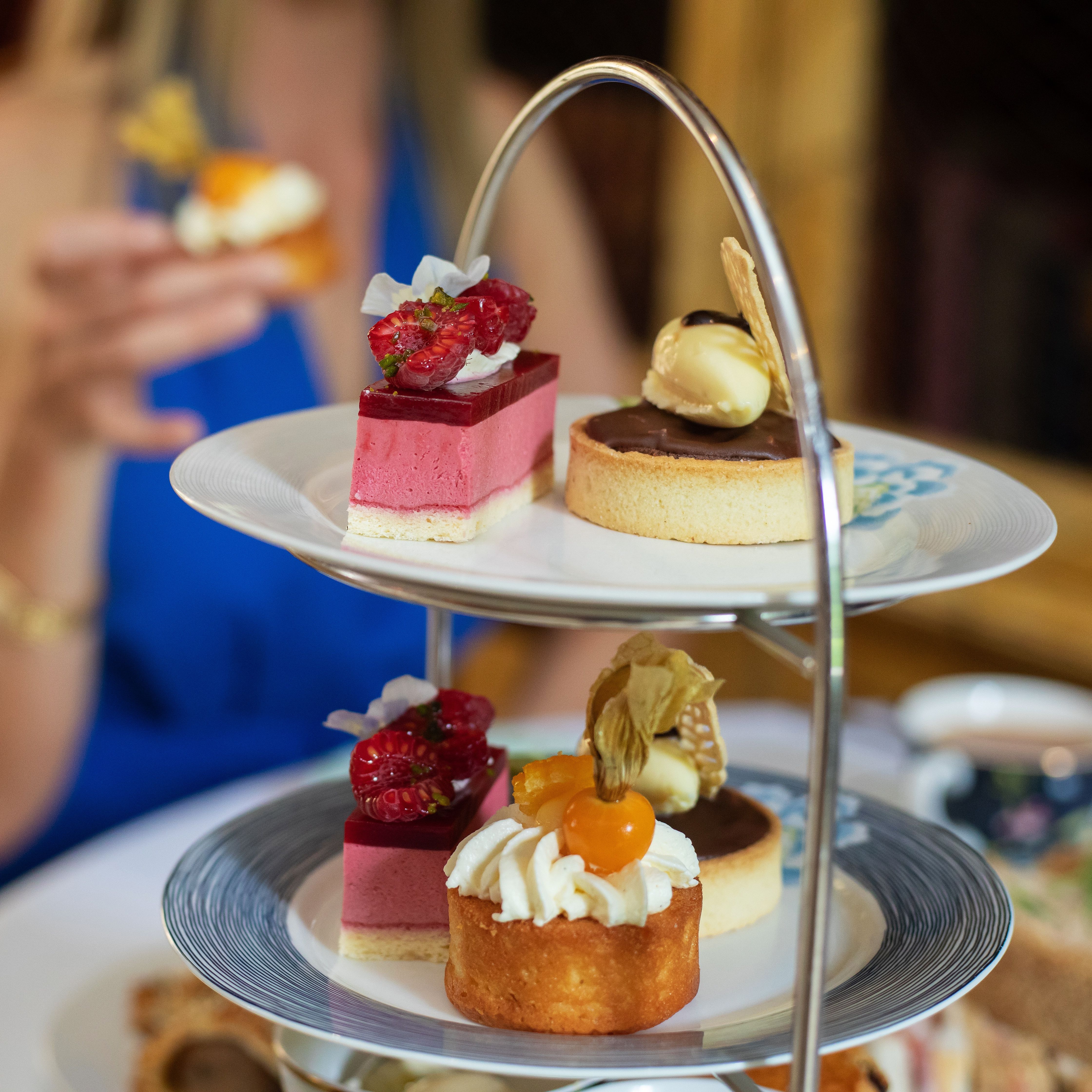 Afternoon tea table with a woman in a blue dress holding a small cake, and a tiered stand filled with colorful desserts and pastries.