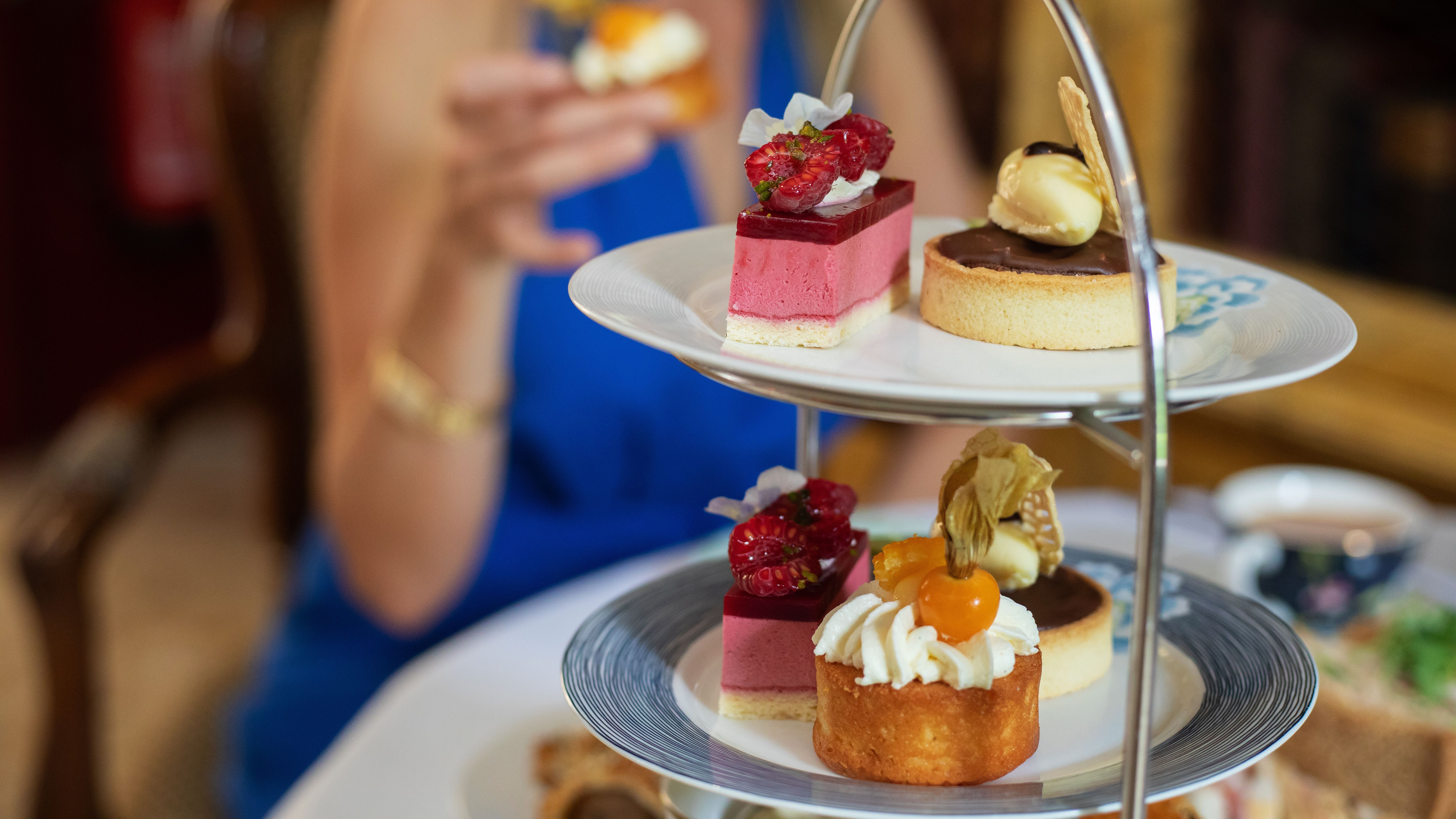 Afternoon tea table with a woman in a blue dress holding a small cake, and a tiered stand filled with colorful desserts and pastries.